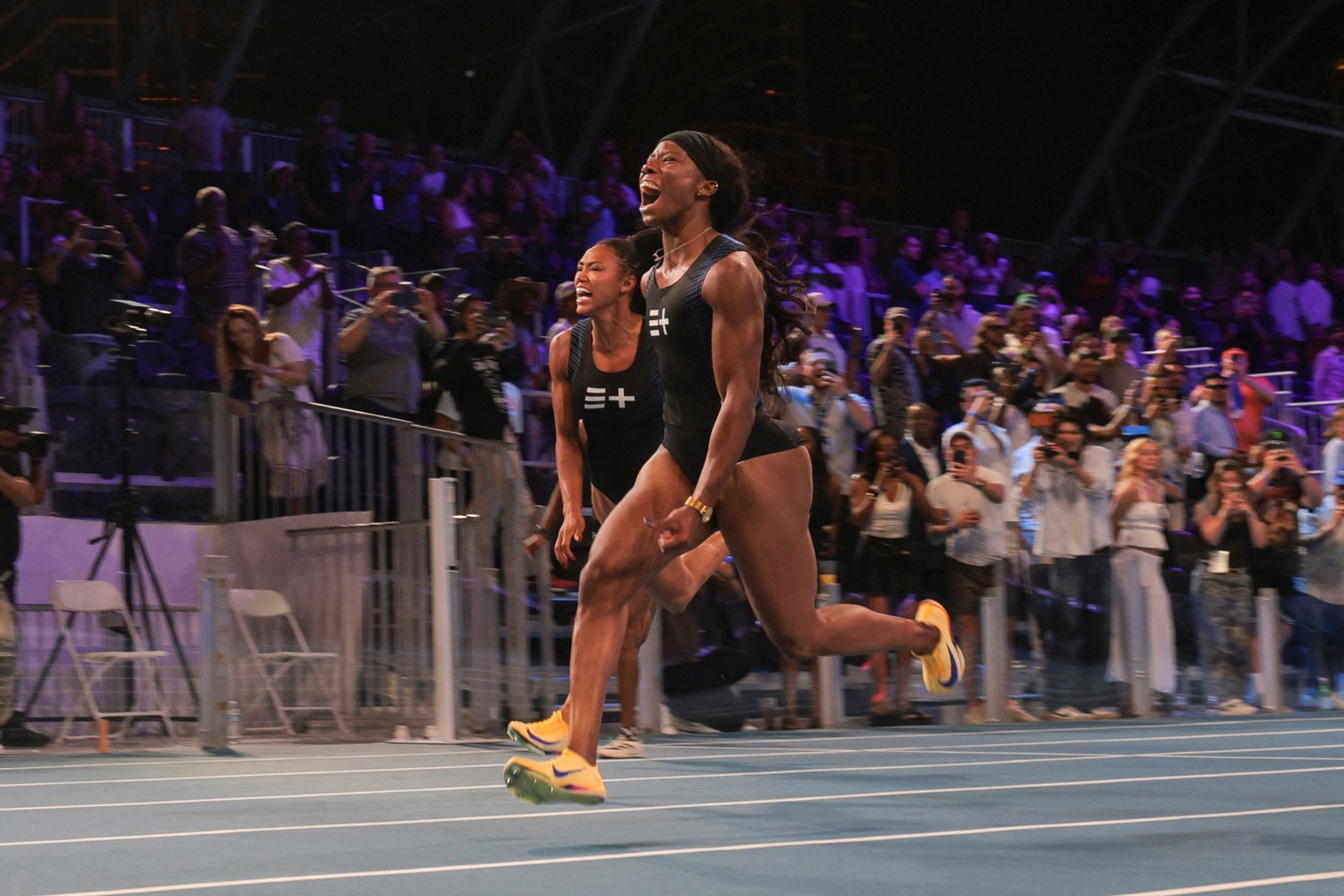Two female athletes sprinting on a blue track, both in black uniforms and yellow shoes, with a crowd cheering wildly in the background.