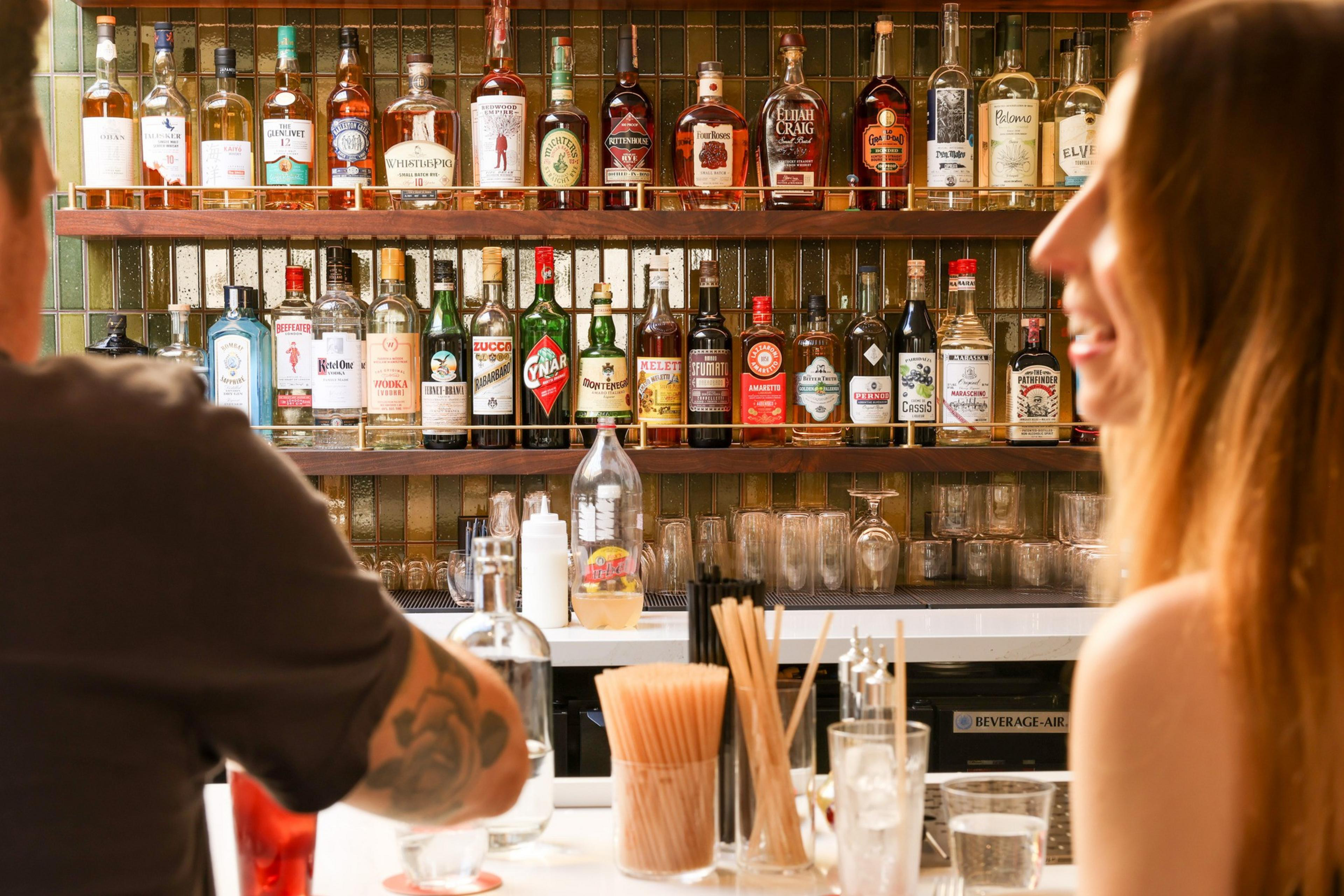 Two people sit at a bar with a variety of liquor bottles on shelves behind them. The bar is stocked with glasses and mixing tools in the foreground.