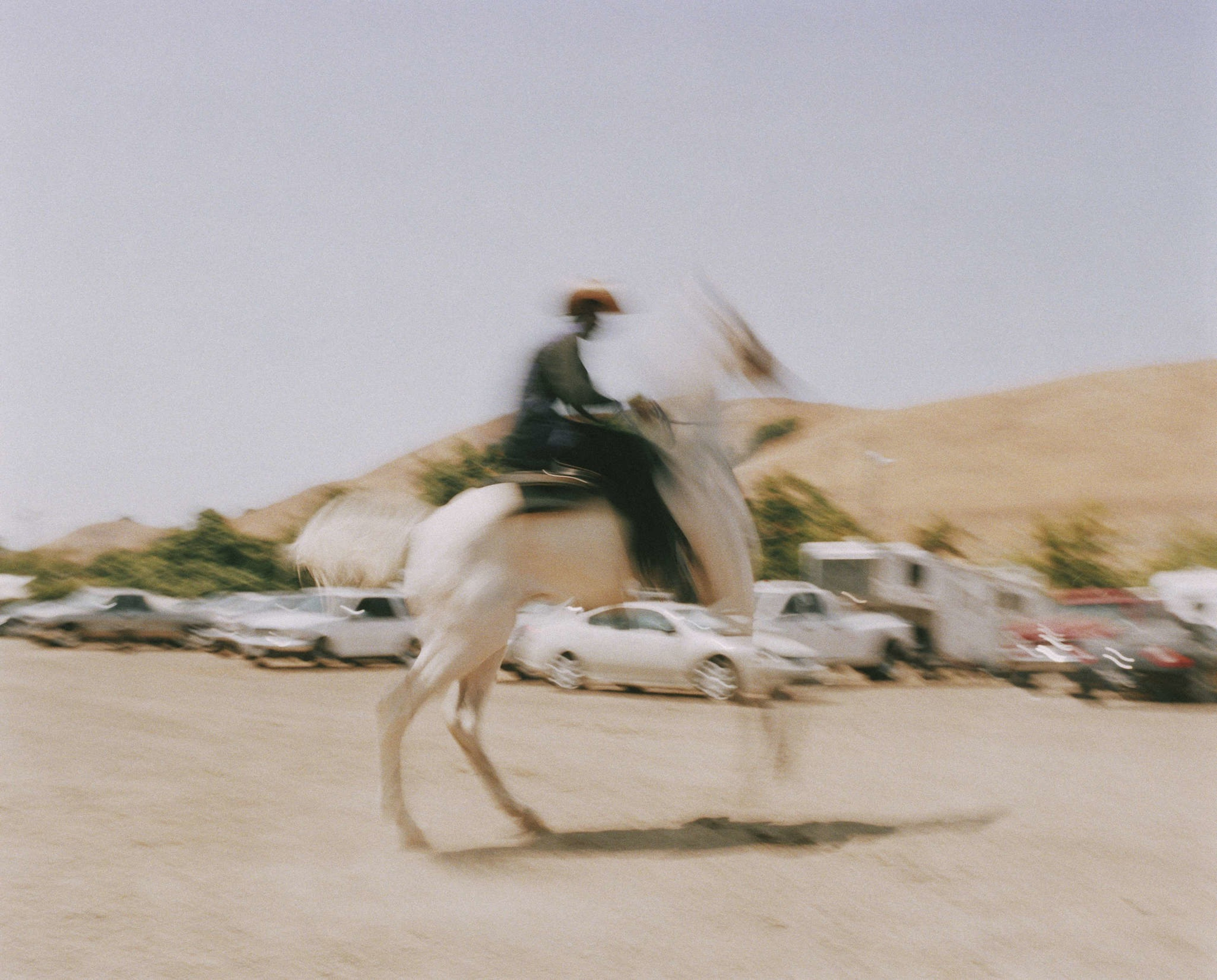 The image shows a blurry figure riding a white horse in motion. The background features parked cars and trailers with a bright, clear sky.