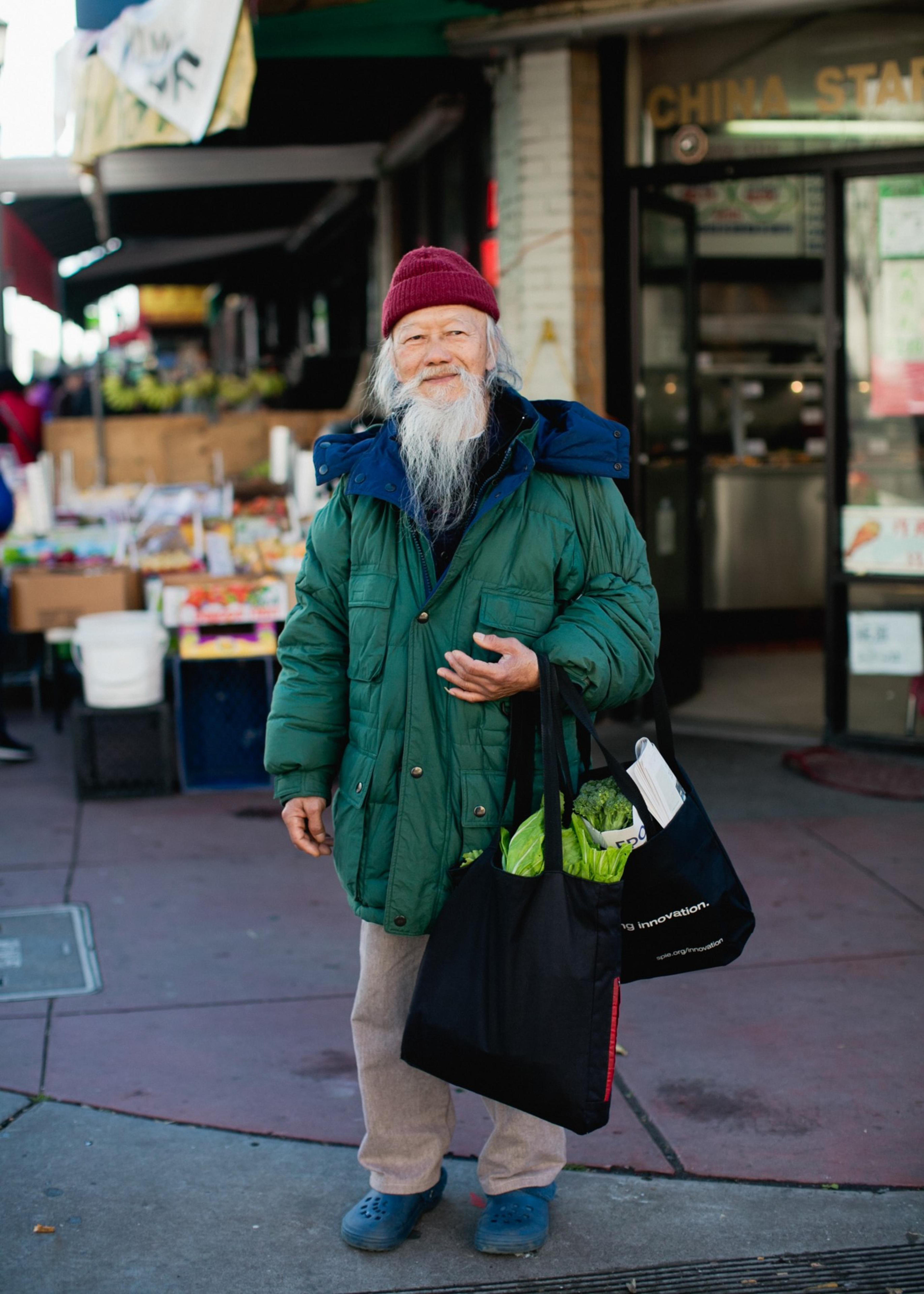 An elderly man with a long white beard wears a red beanie, green puffy jacket, beige pants, and blue shoes, holding a black reusable bag with groceries.