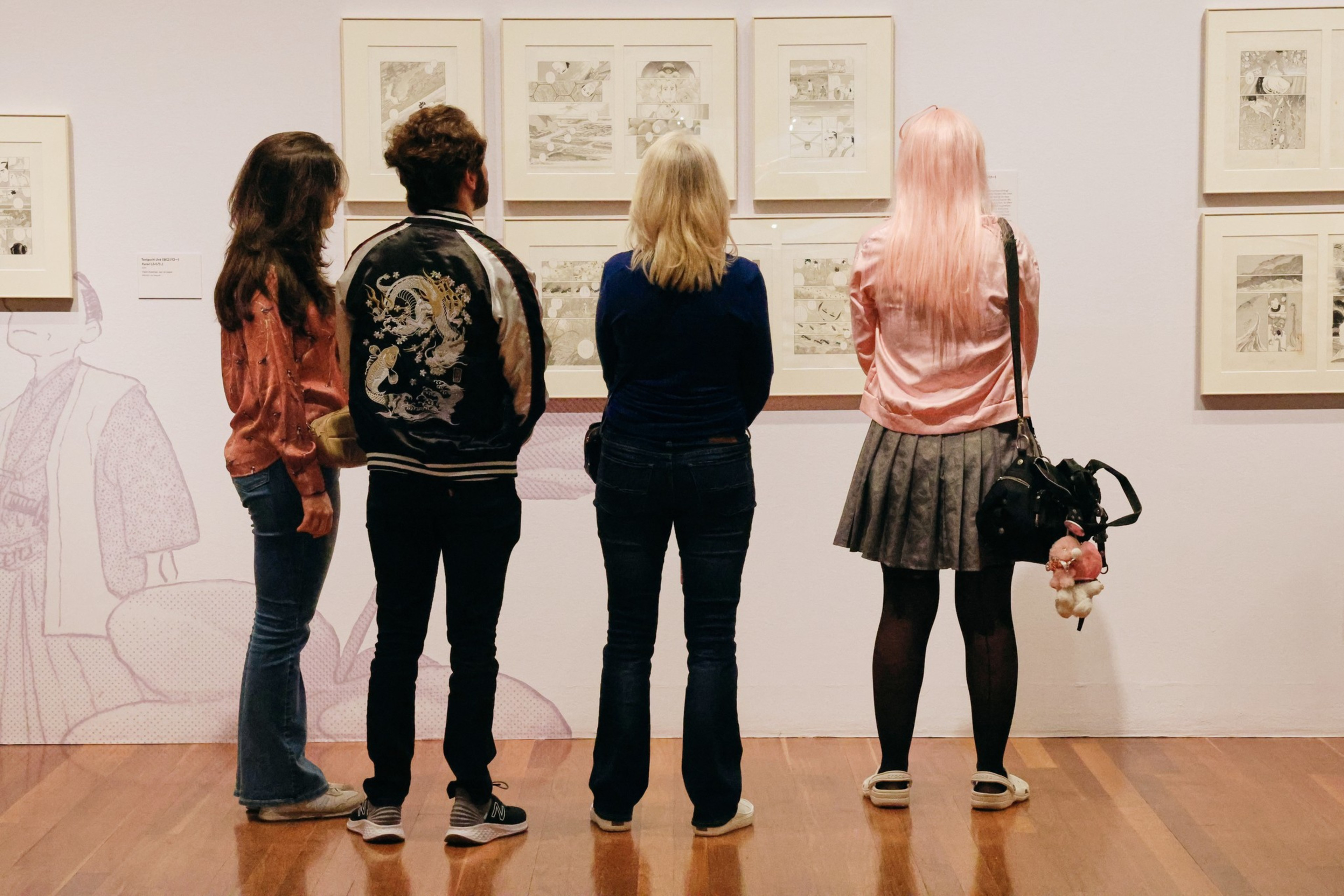 Four people stand side by side, viewing framed comic-style artwork on a museum wall, with wooden floors and soft lighting.