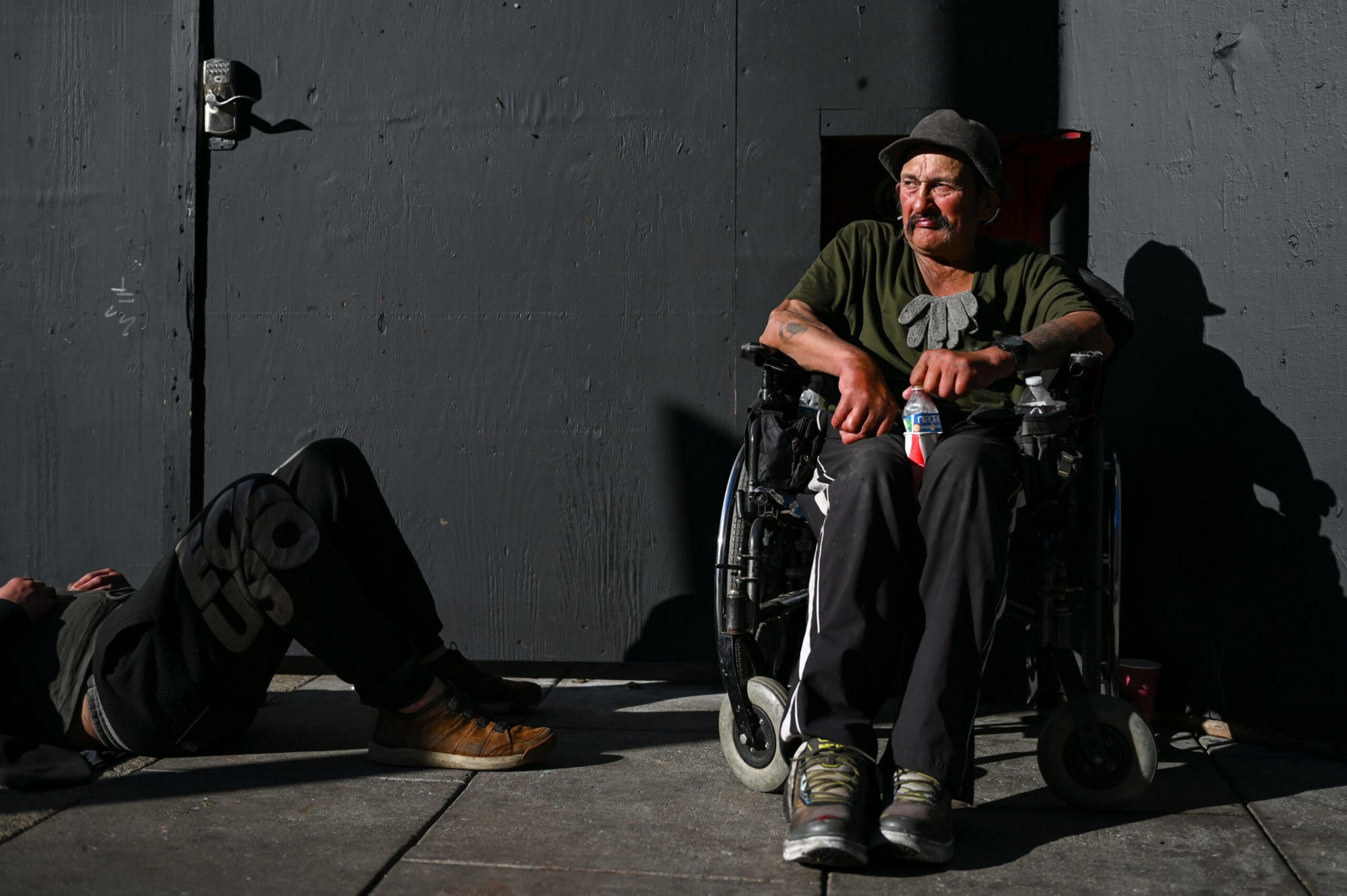 A man sits in a wheelchair holding a water bottle, wearing a hat and gloves, while another person lies on the ground nearby against a dark wall.