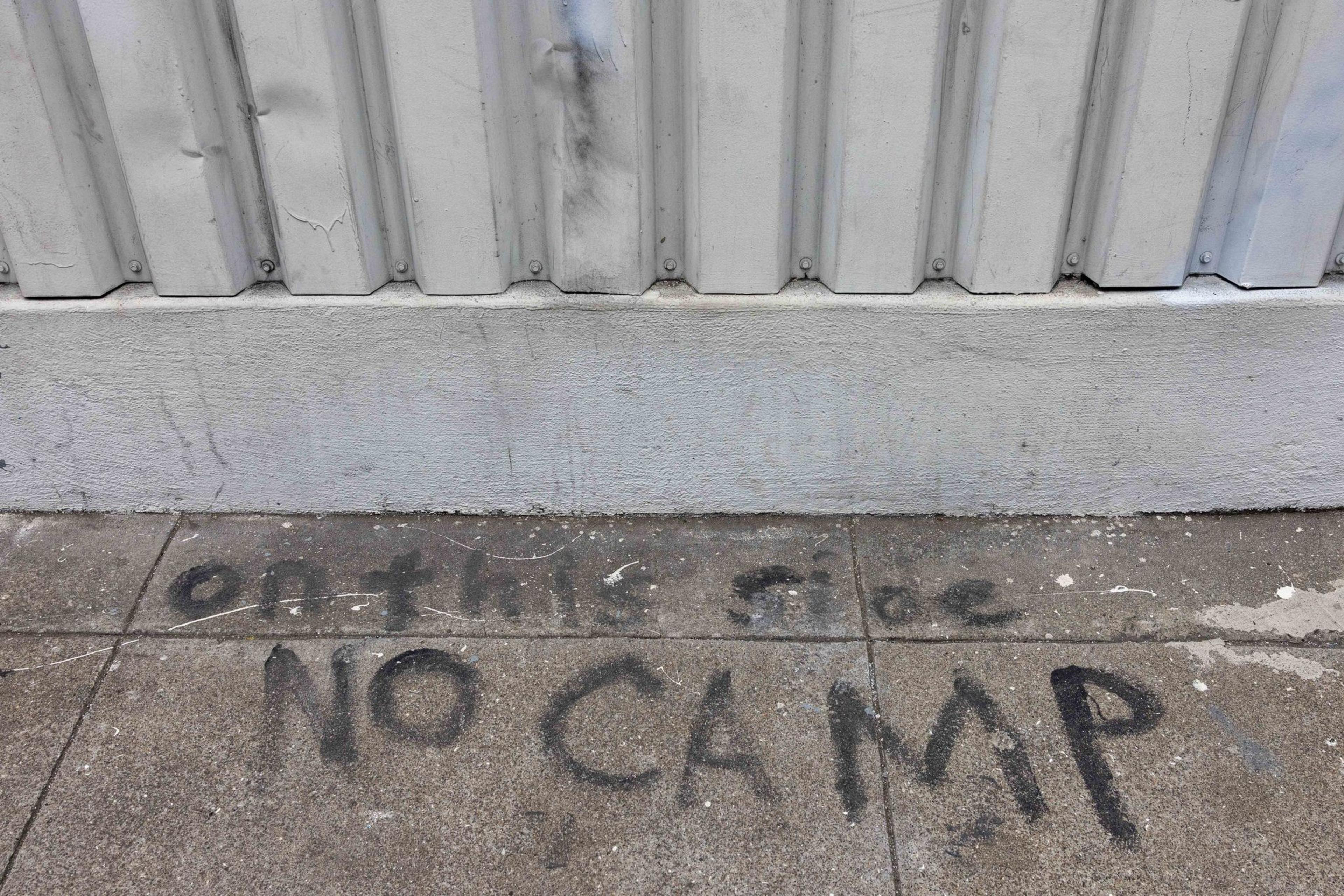 A gray concrete wall and sidewalk feature handwritten text reading &quot;on this side NO CAMP&quot; in black paint.