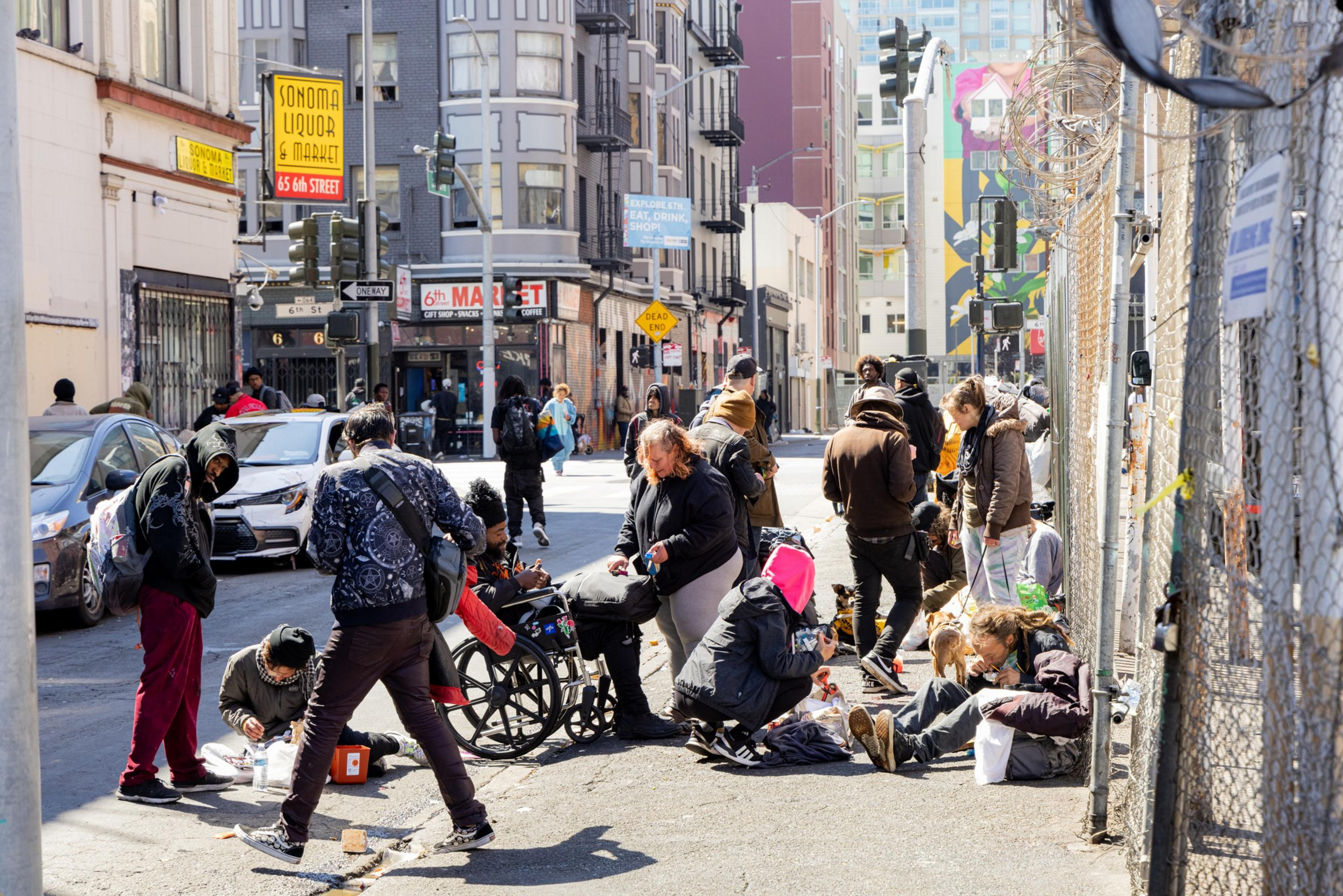 A group of people, some sitting and others standing, gather on a street corner near a liquor store and market, surrounded by buildings and parked cars.