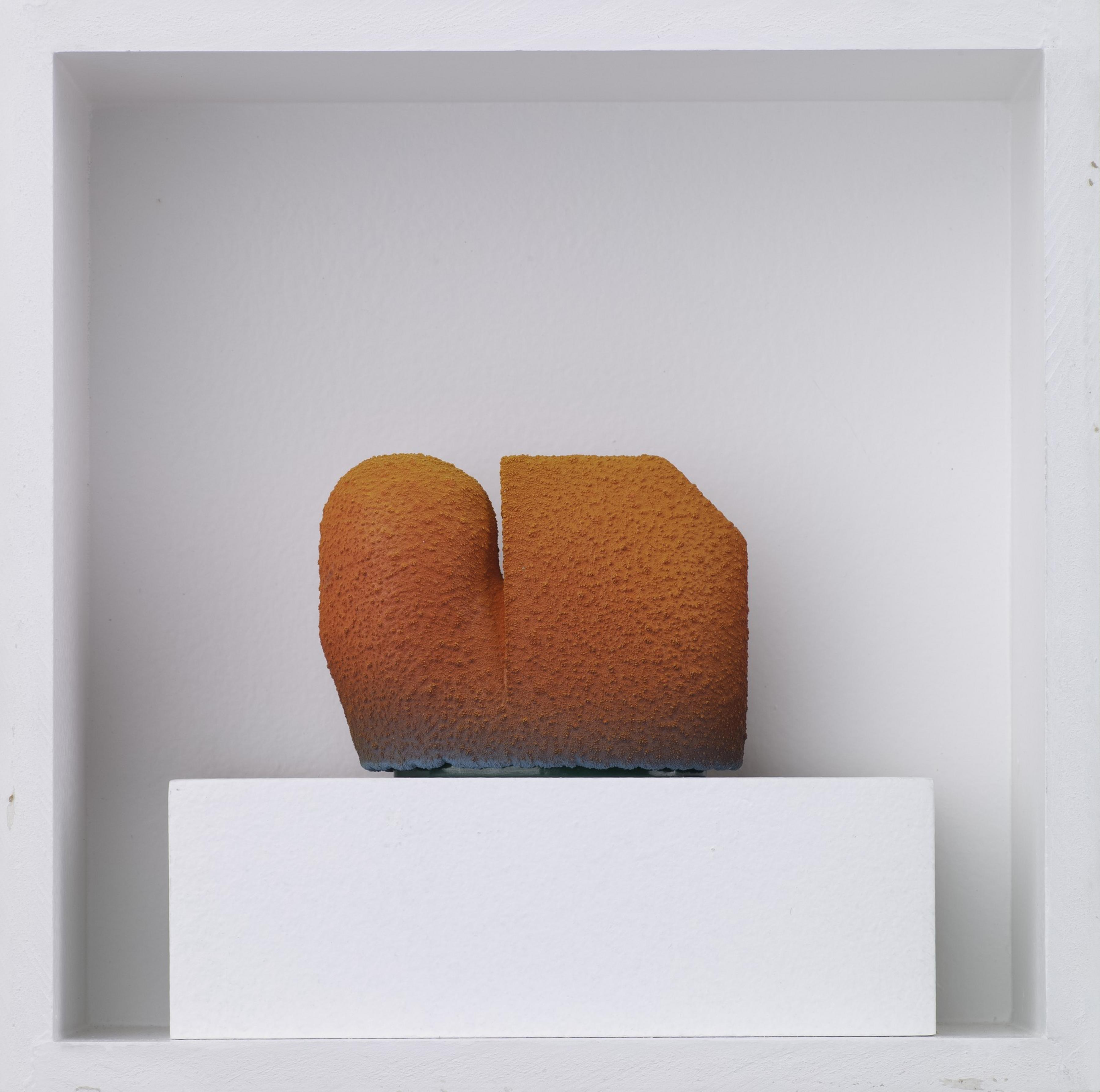 A textured, gradient orange sculpture with a rounded left side and an angular right side sits on a white pedestal inside a recessed white wall niche.