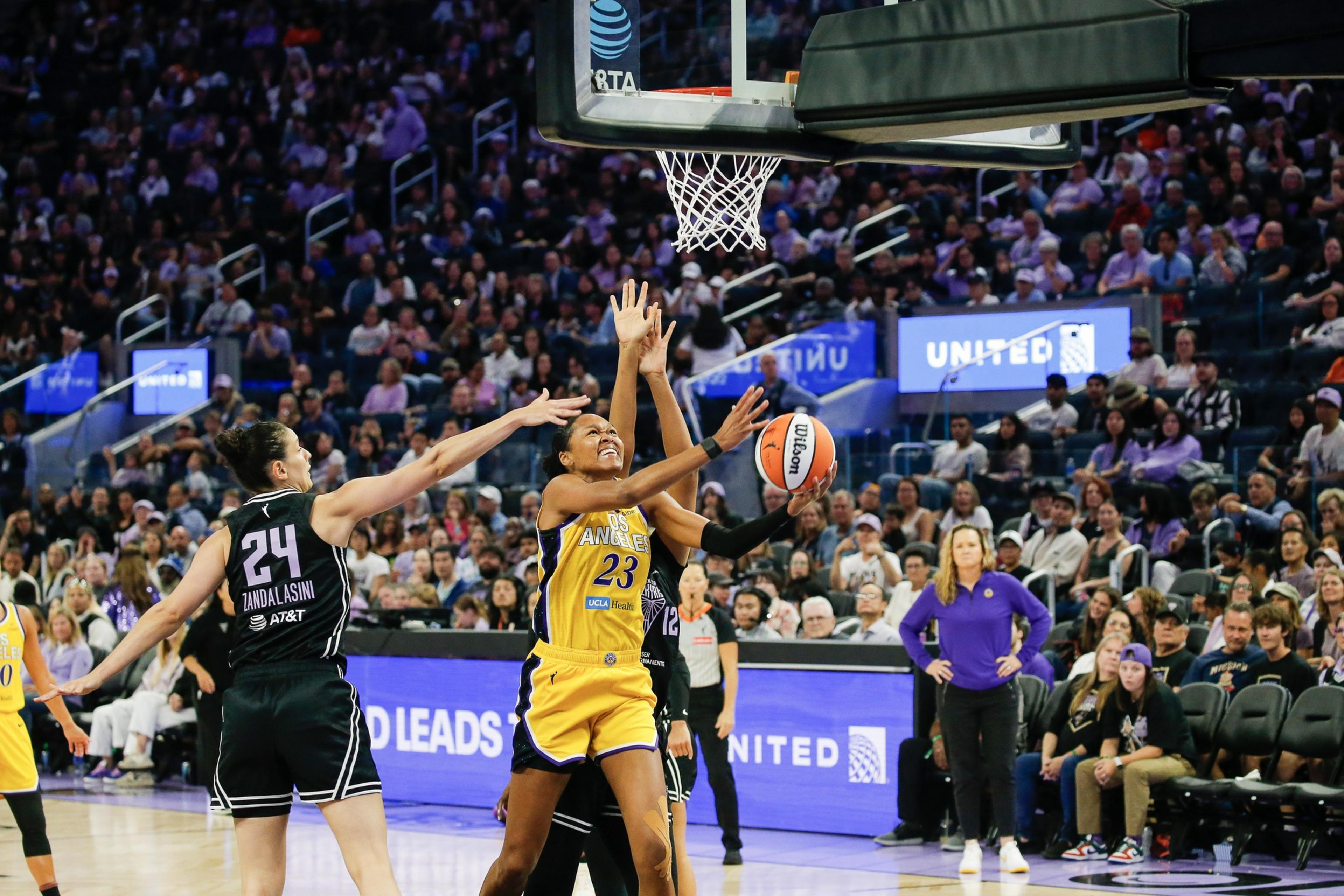 A basketball player in a yellow uniform leaps to shoot while two defenders in black try to block her under the hoop with a crowded arena in the background.