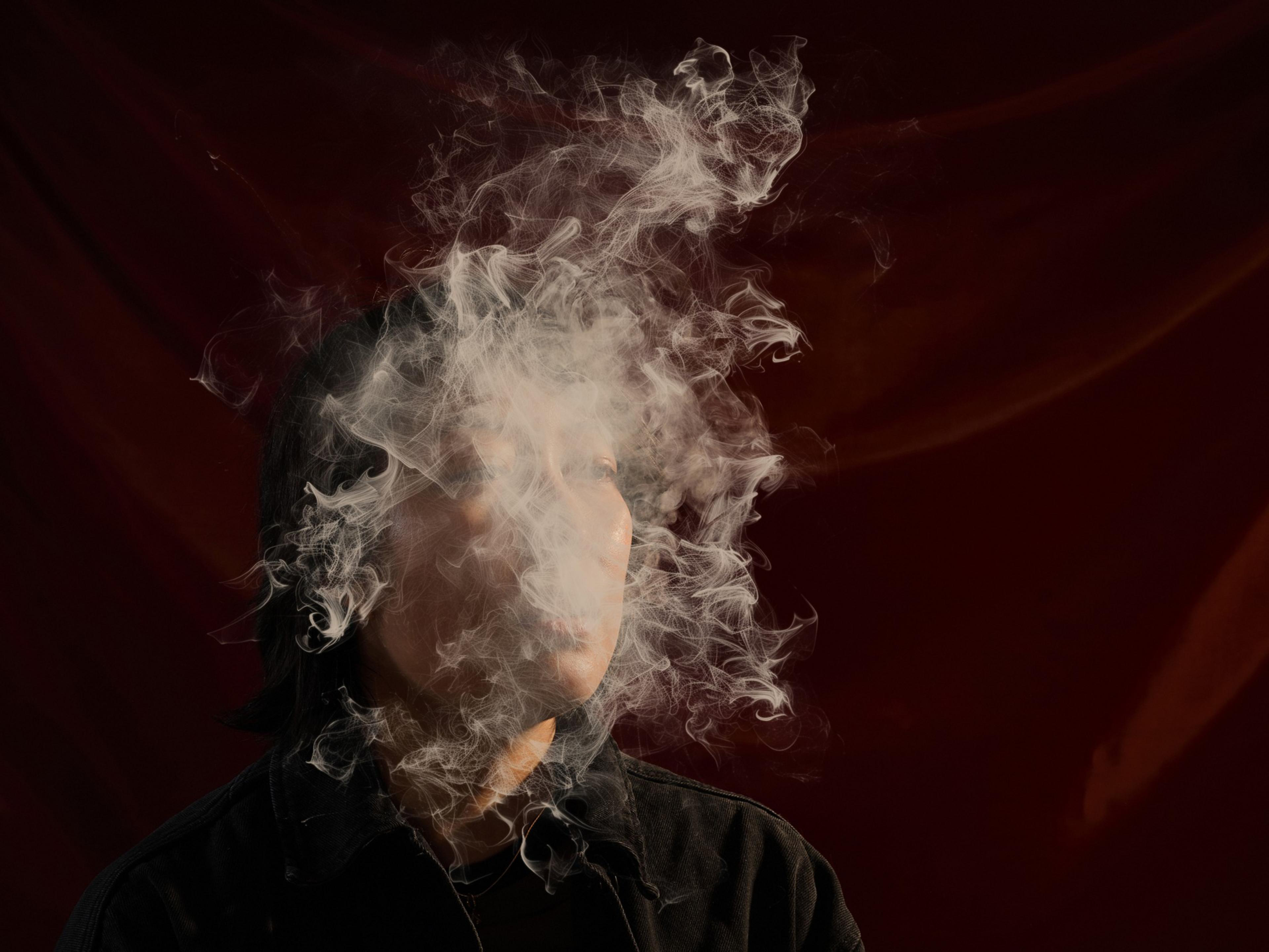 A person is partially obscured by swirling white smoke against a dark red background, creating a mysterious and moody atmosphere.