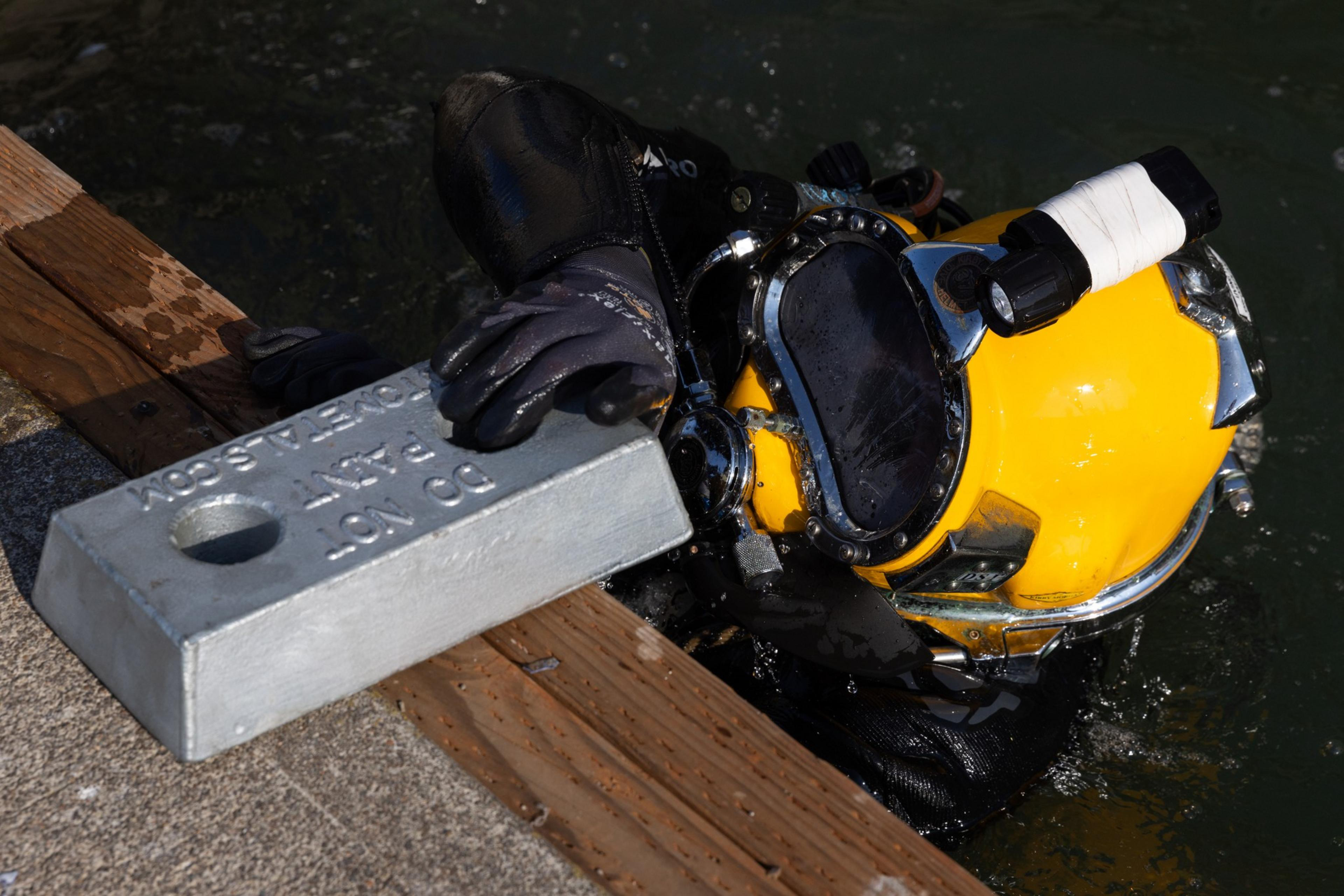 A diver in a yellow helmet and black gloves holds a 30-pound metal weight near a wooden dock edge above murky water.