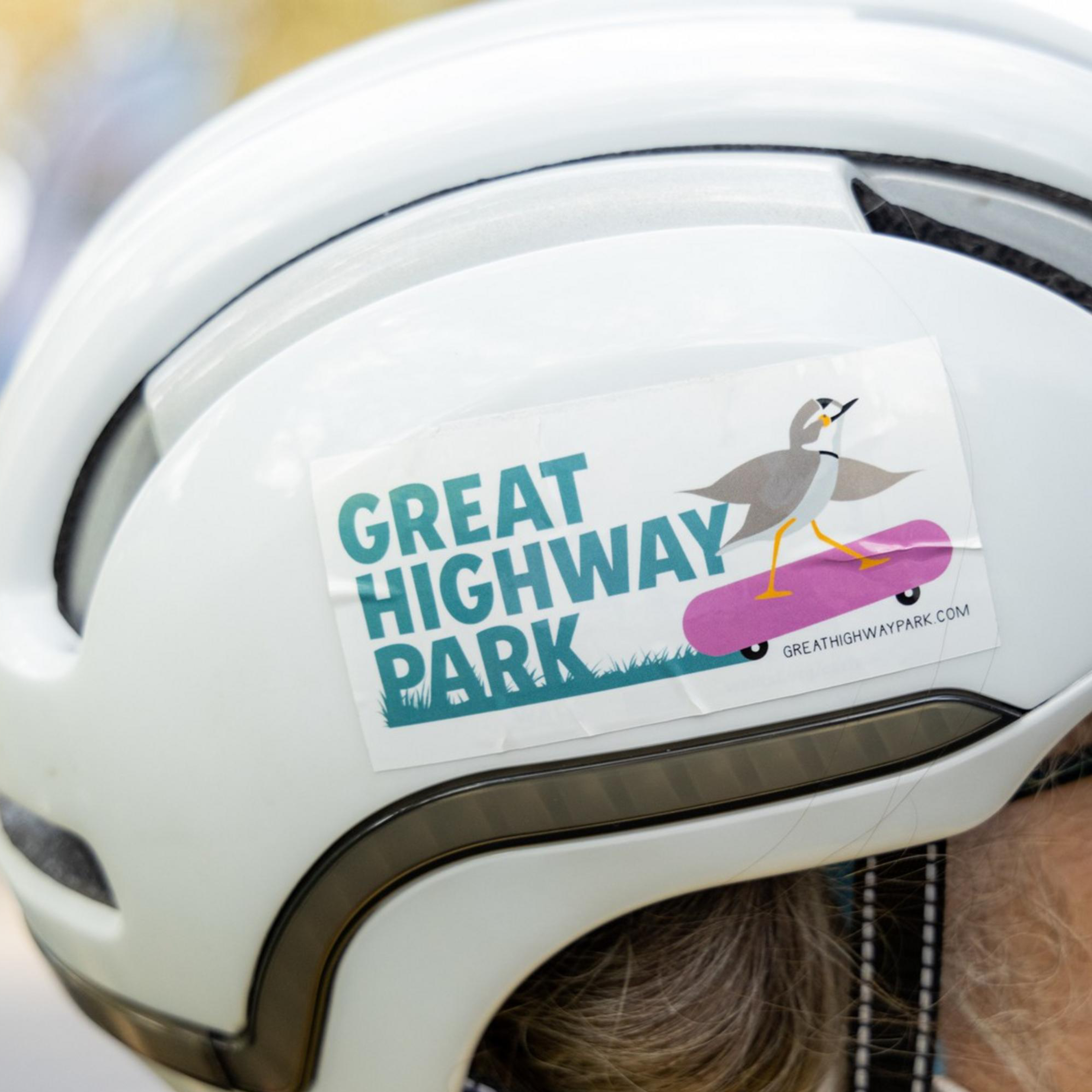 An individual wearing a white helmet with a sticker that reads &quot;Great Highway Park&quot; and depicts a bird on a skateboard.