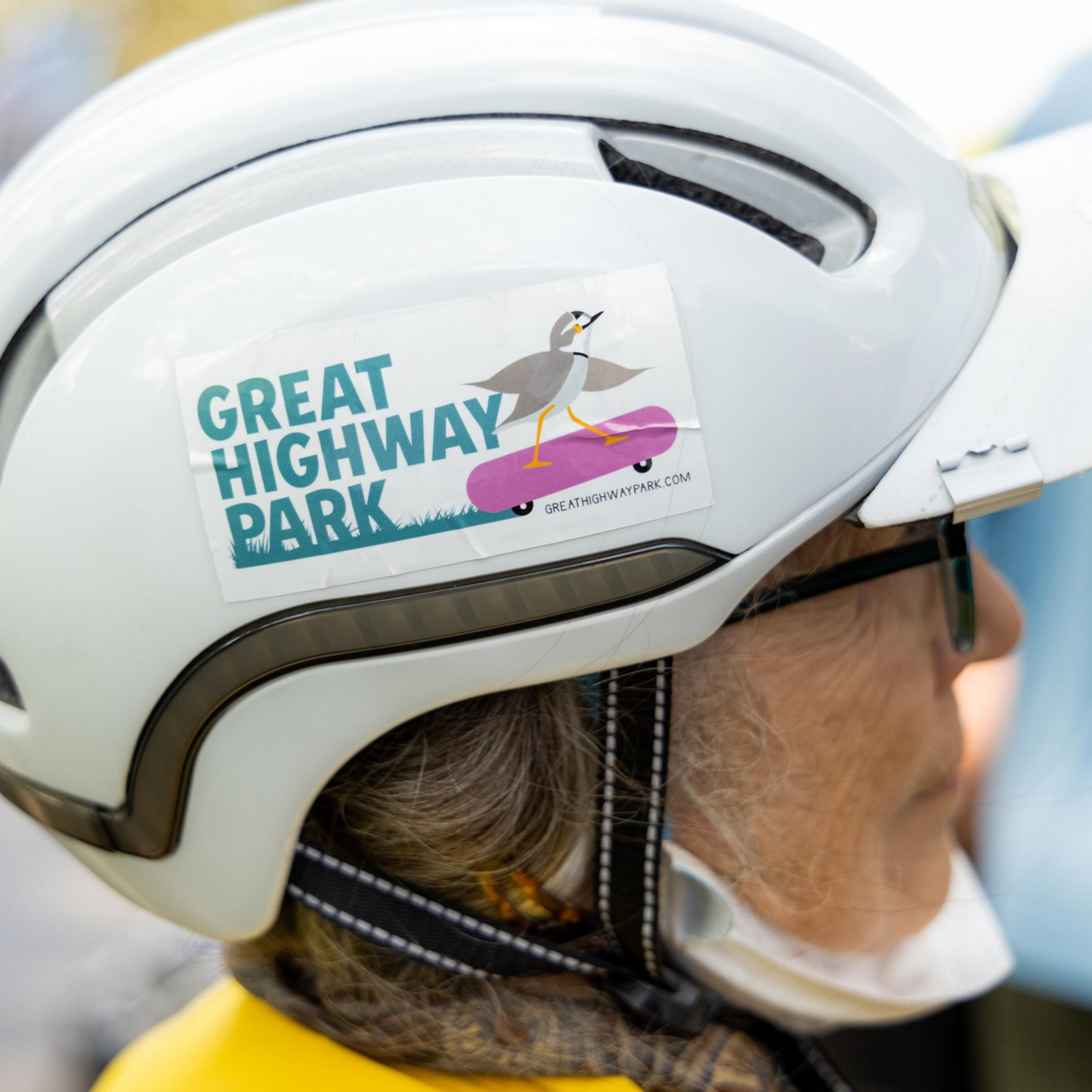 An individual wearing a white helmet with a sticker that reads &quot;Great Highway Park&quot; and depicts a bird on a skateboard.
