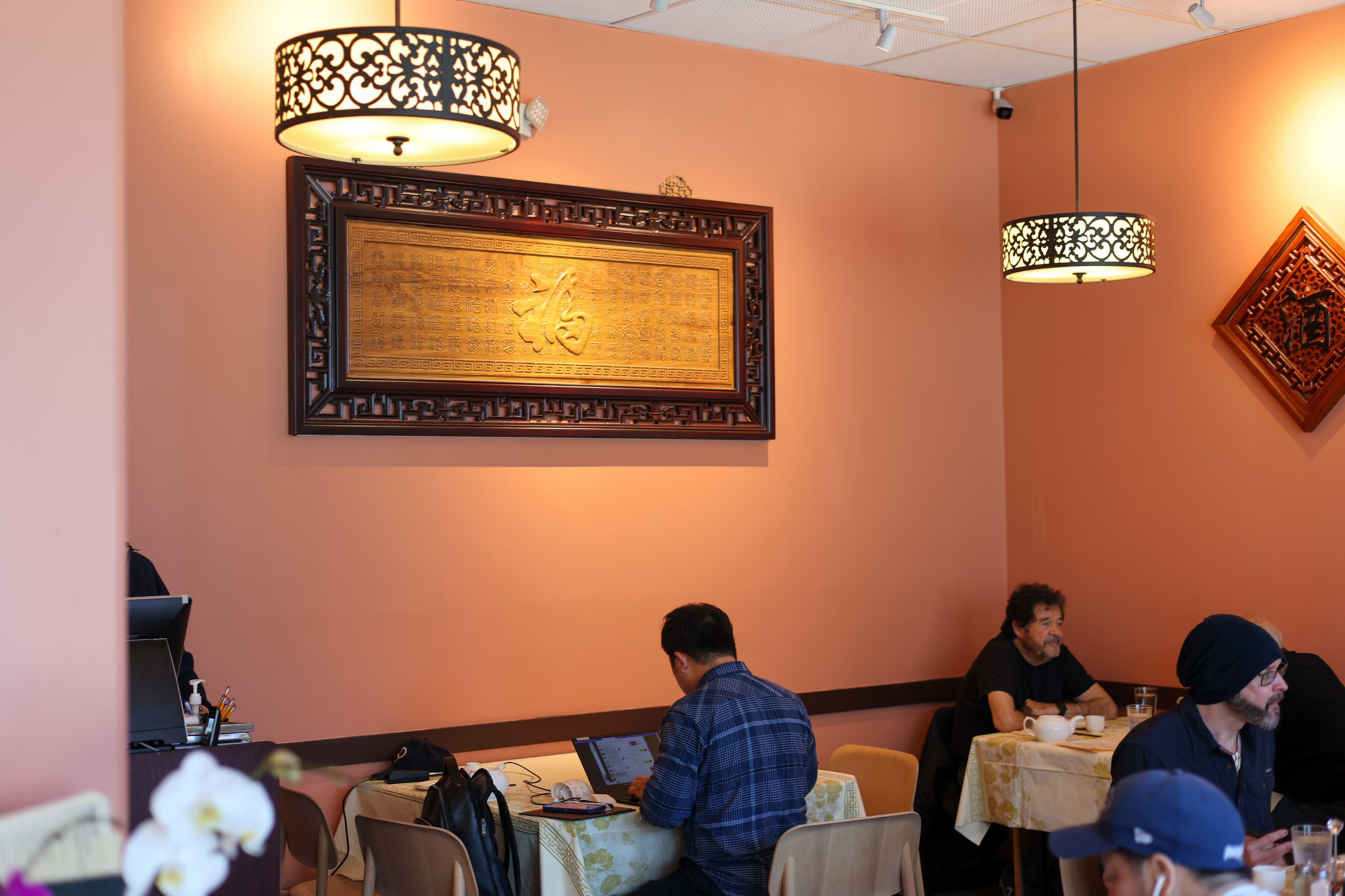 People sit at tables with laptops and food in a warmly lit cafe featuring peach walls, hanging lantern-style lights, and framed Asian-style wooden art.