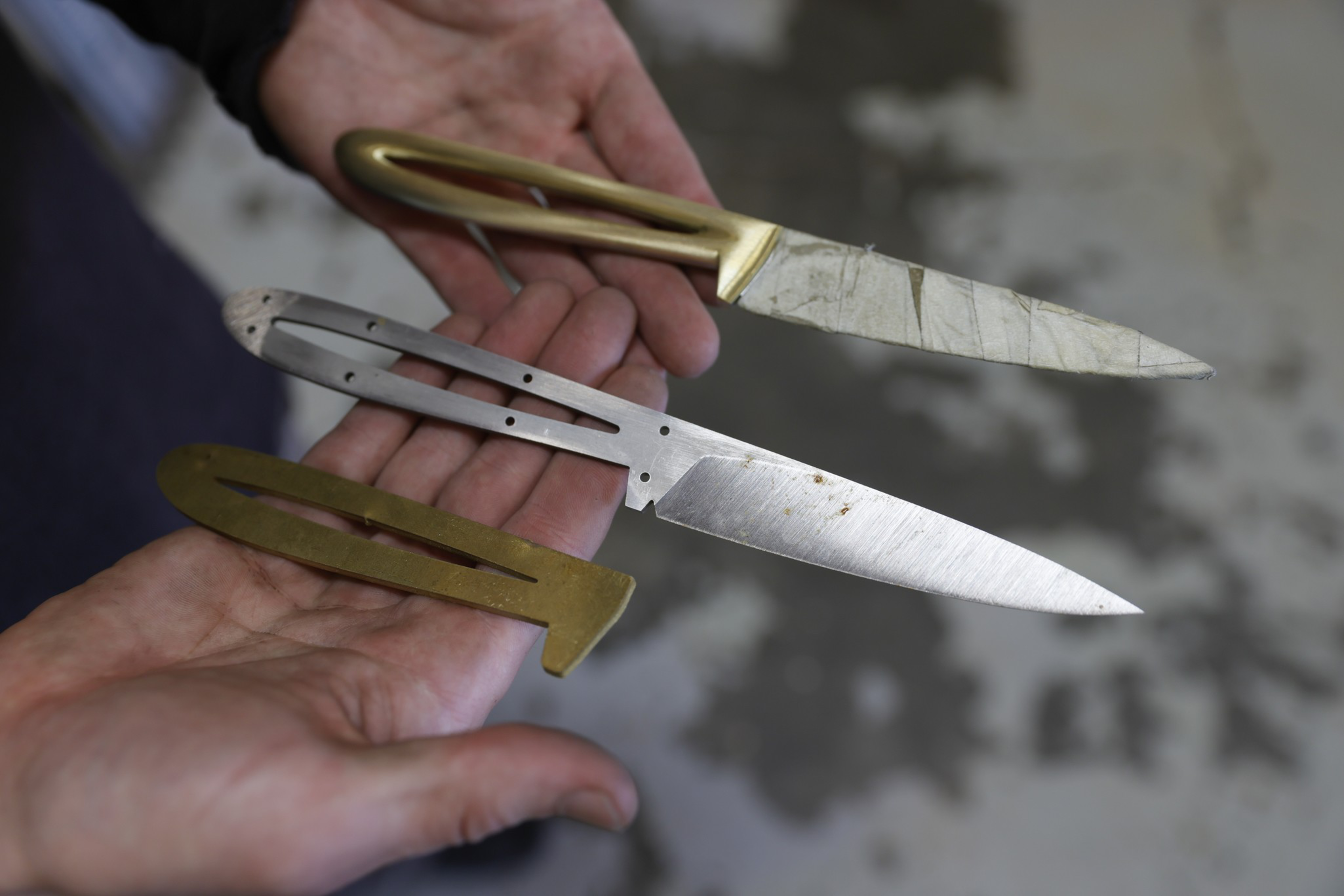 Two hands hold two knives with skeletal handles and one separate gold-colored handle piece, all against a blurred gray background.