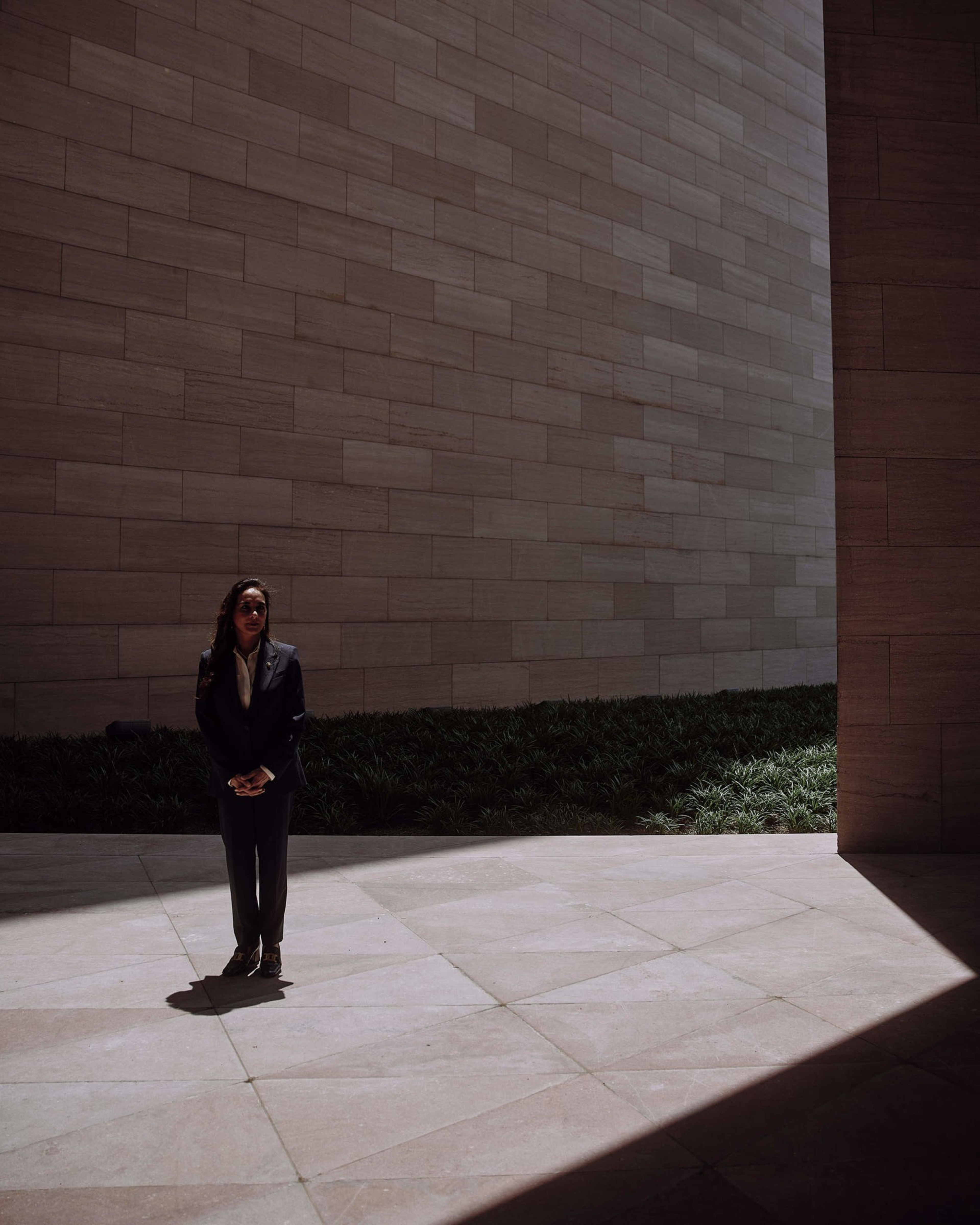 A person in a dark suit stands in a sunlit courtyard, surrounded by large tiled walls and patches of greenery in the background.
