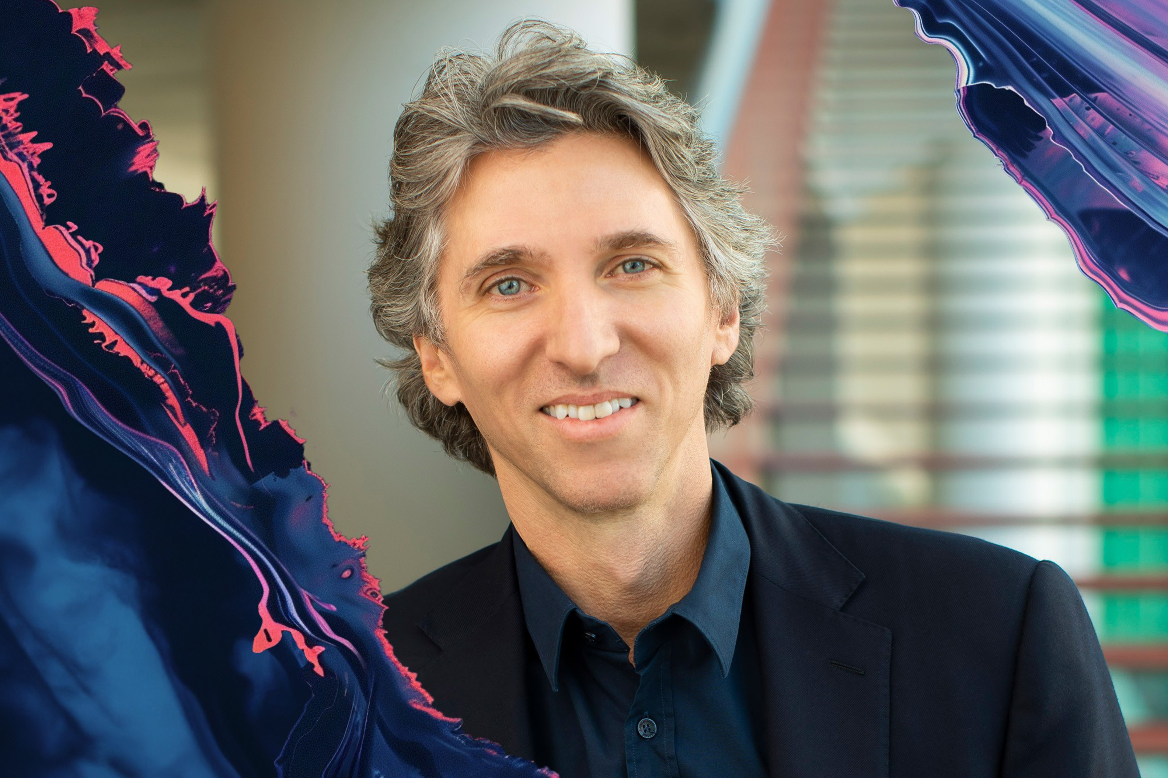 Damian Woetzel, with gray hair, wearing a dark suit and blue shirt, smiles against a blurred background. The image has colorful abstract shapes in the corners.