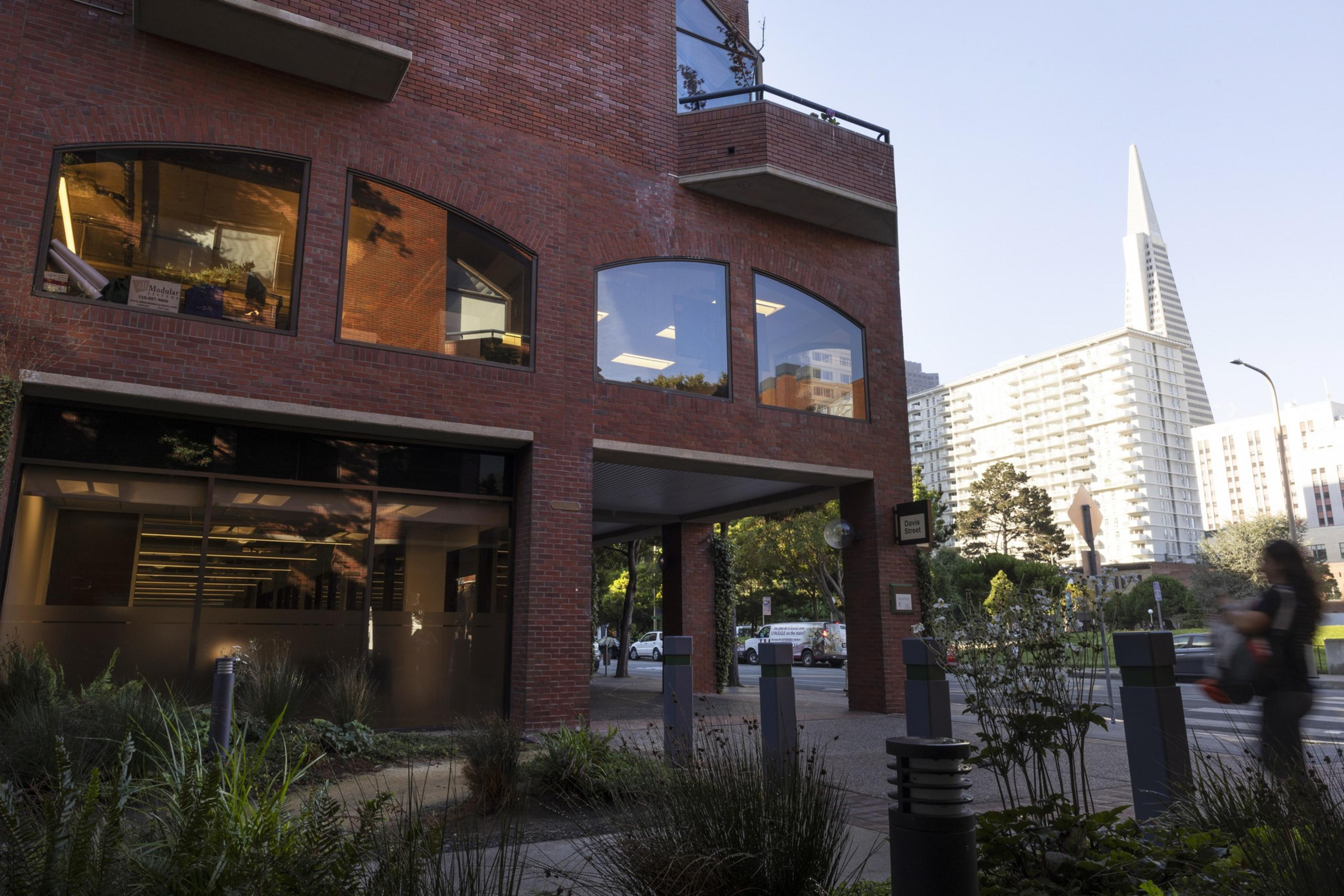 A red brick building with large windows reflects indoor lighting, while a person walks on the sidewalk near plants and modern street fixtures.