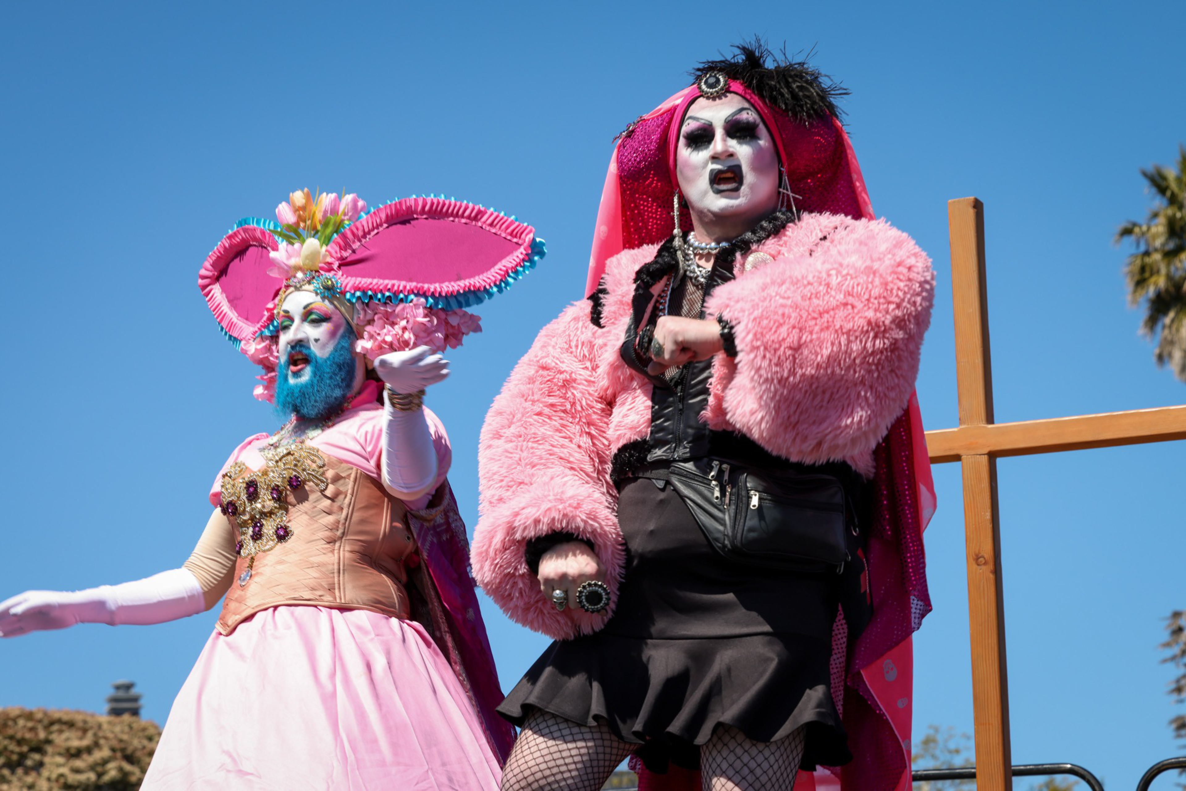 Two performers in vibrant drag costumes pose dramatically; one wears a large pink bunny ear headpiece, the other a fluffy pink jacket and black skirt.