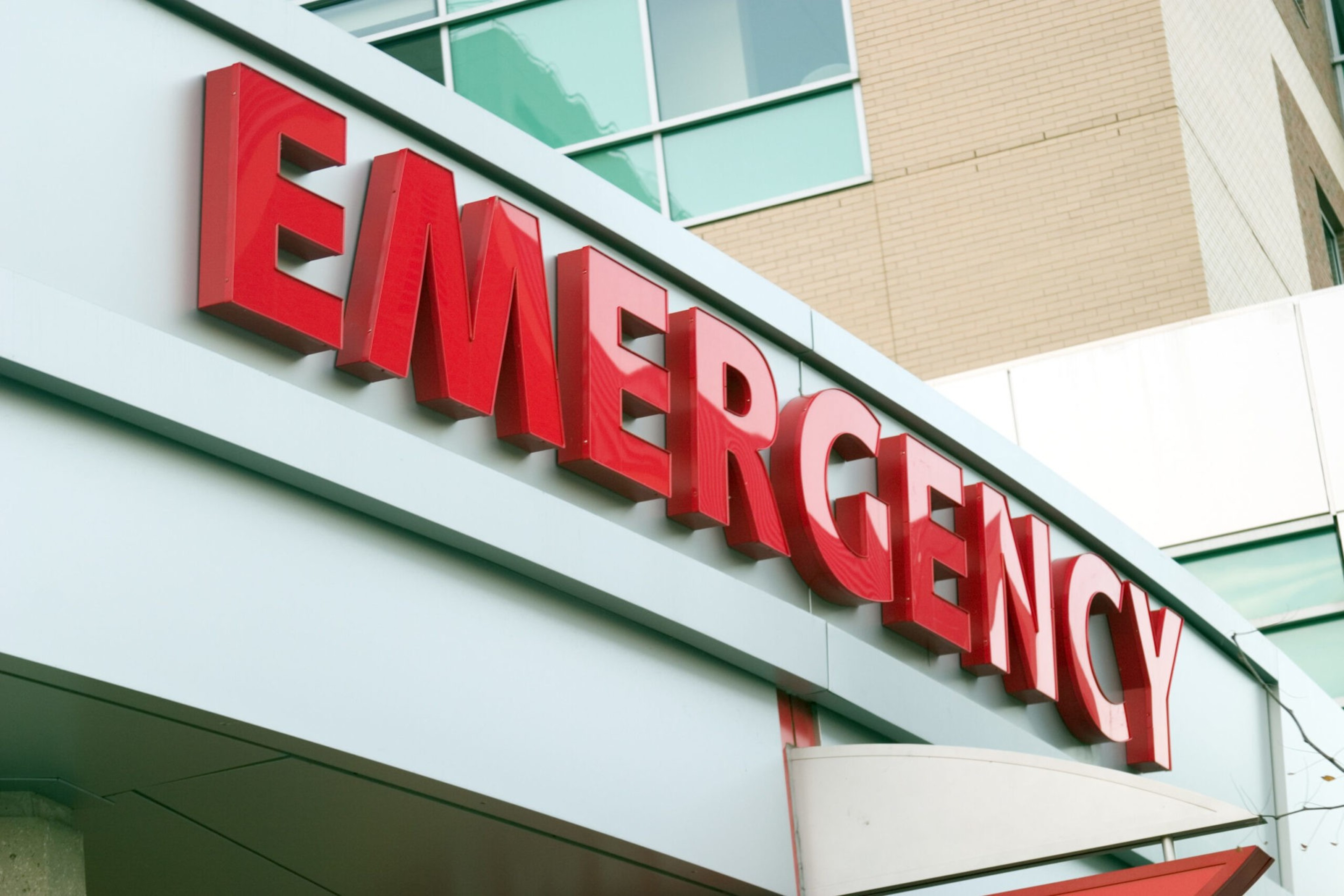 A large red "EMERGENCY" sign is mounted above the entrance of a hospital building with glass windows and beige brick walls.