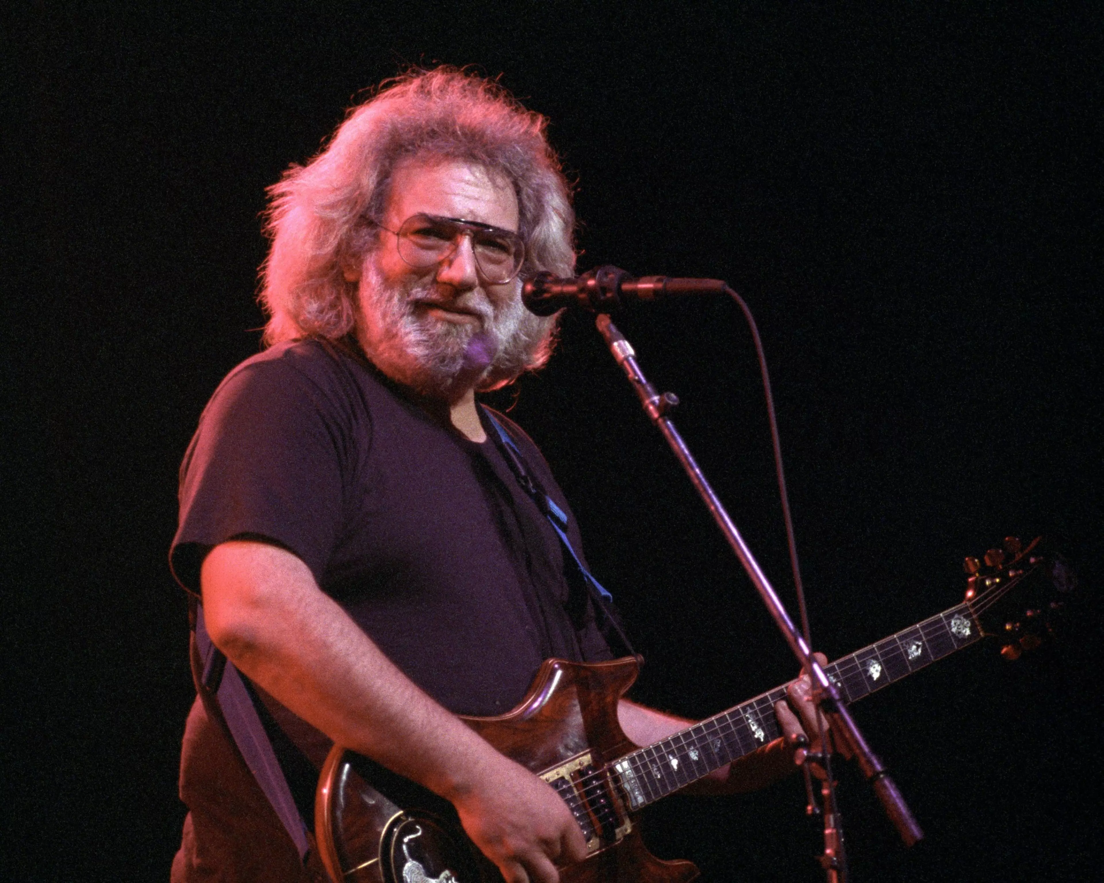 A bearded man with glasses plays an electric guitar on stage, smiling towards the audience. The background is dark, highlighting him under warm lighting.