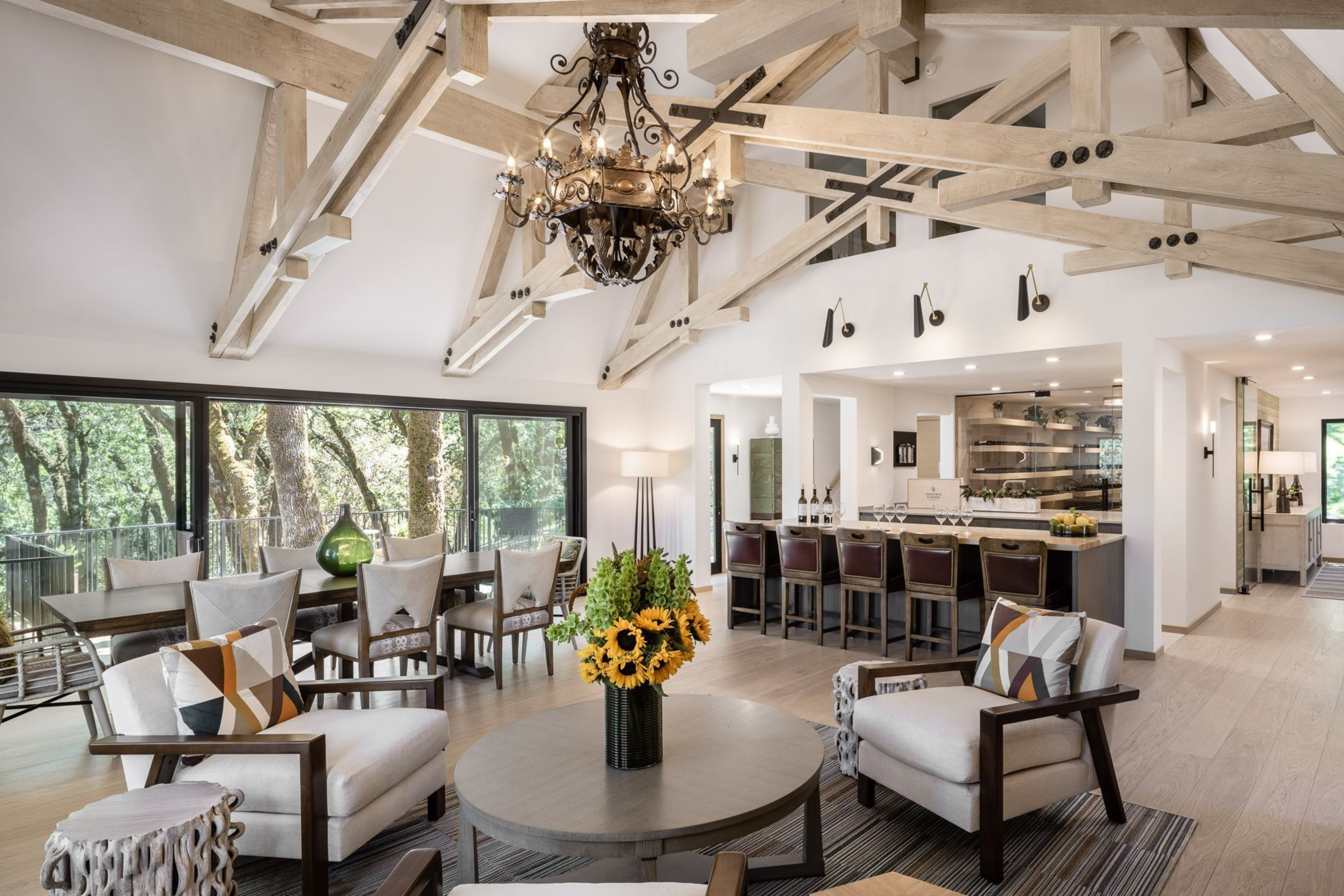 A spacious living and dining area with exposed wooden beams, a chandelier, large windows, sunflowers on a round table, and a bar with stools.