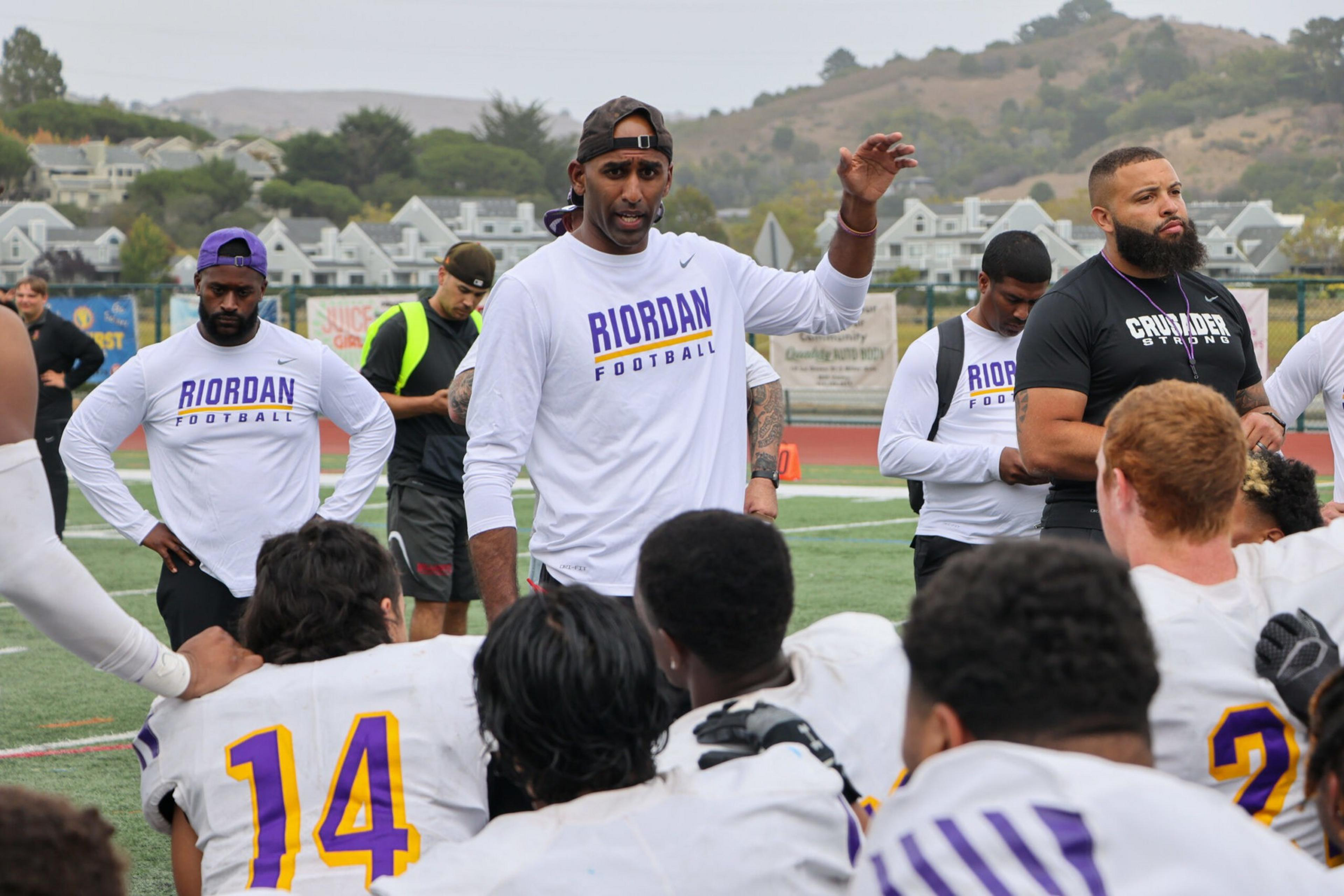 A coach wearing a "Riordan Football" shirt talks to a seated football team on a field, with other coaches and players standing nearby.