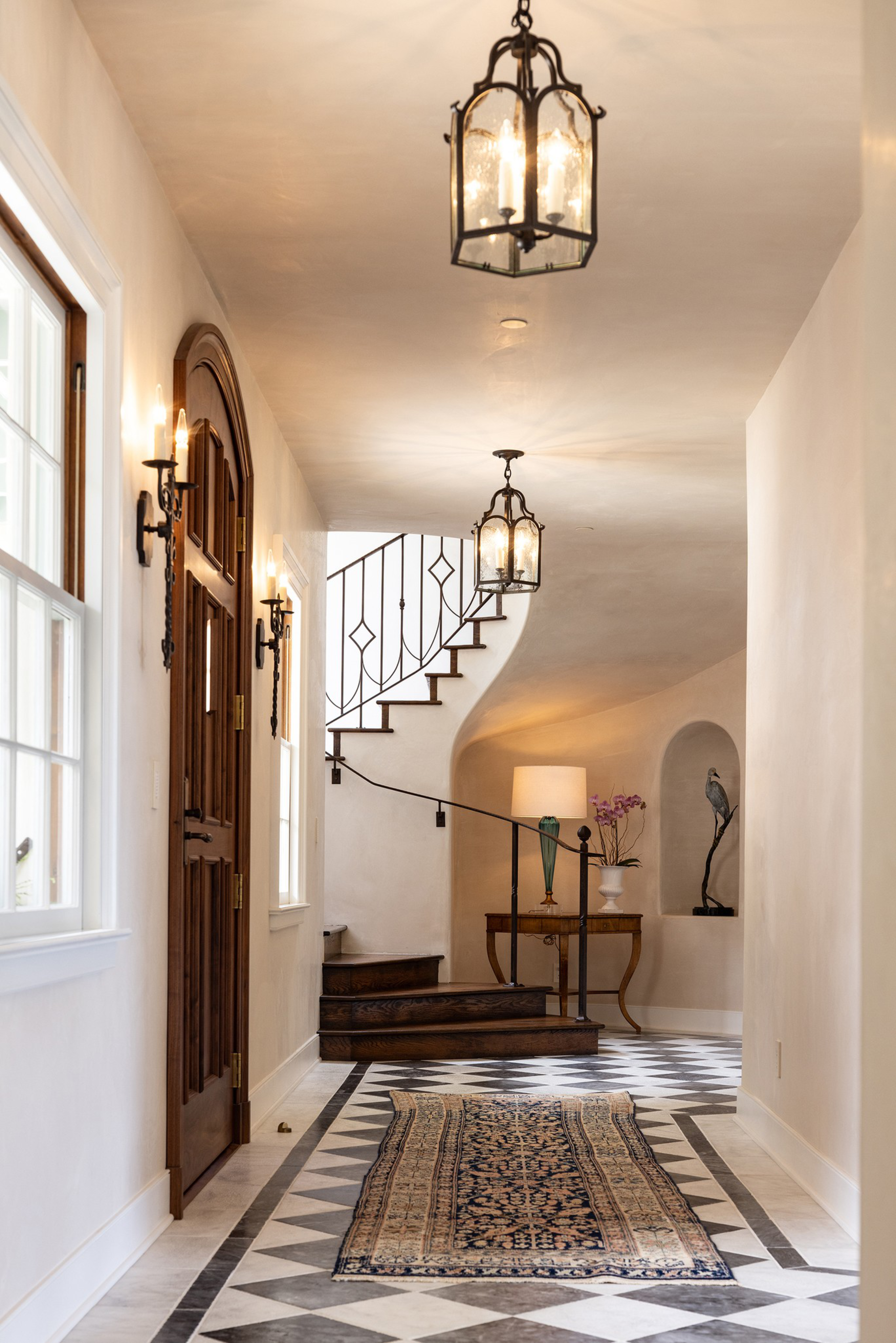 A hallway with a dark wooden door, patterned floor tiles, an ornate rug, curved wooden stairs with a black railing, and a table with a lamp and sculpture.