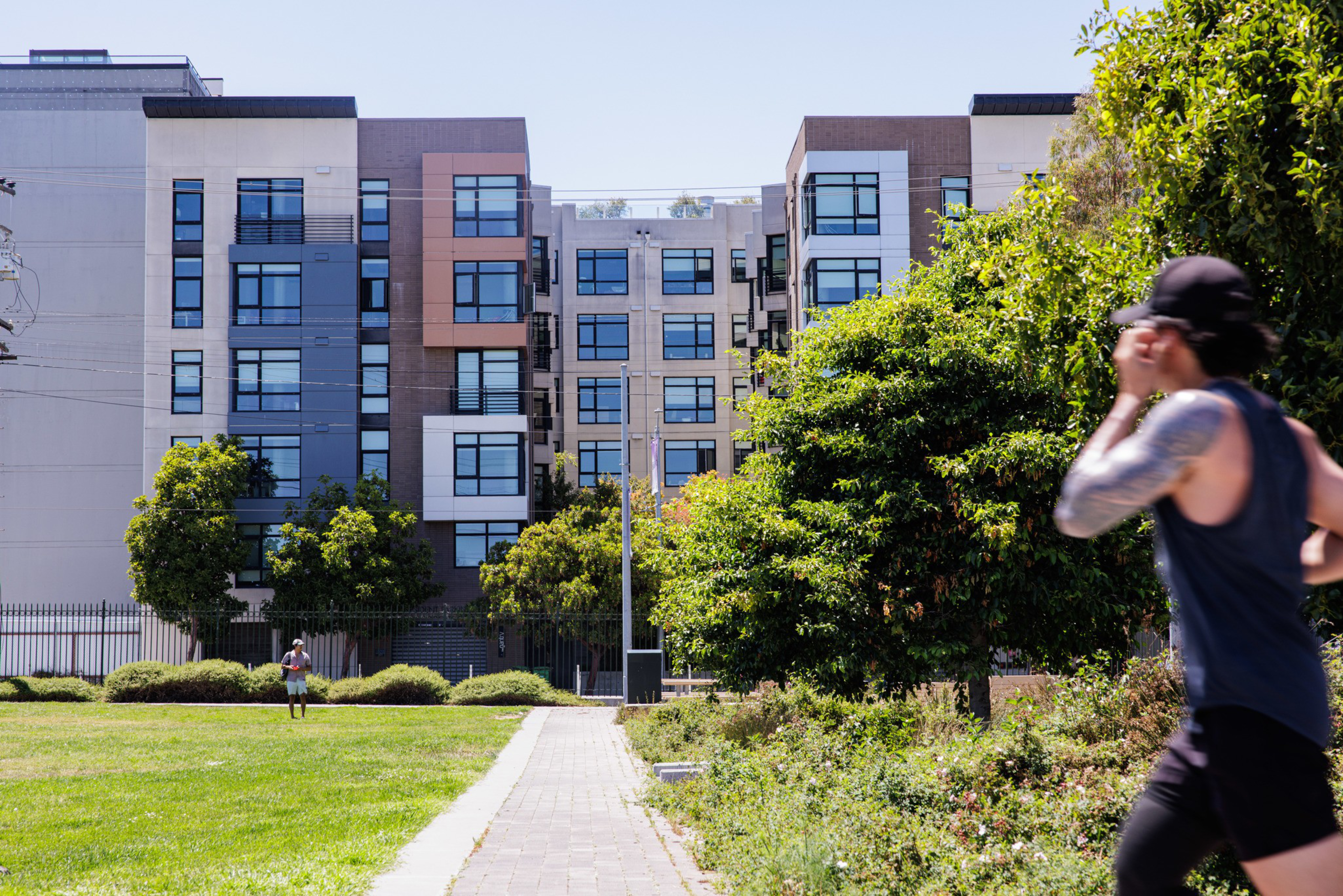 A modern apartment building stands behind a grassy park where a person jogs on a path and another walks near trees and bushes on a sunny day.