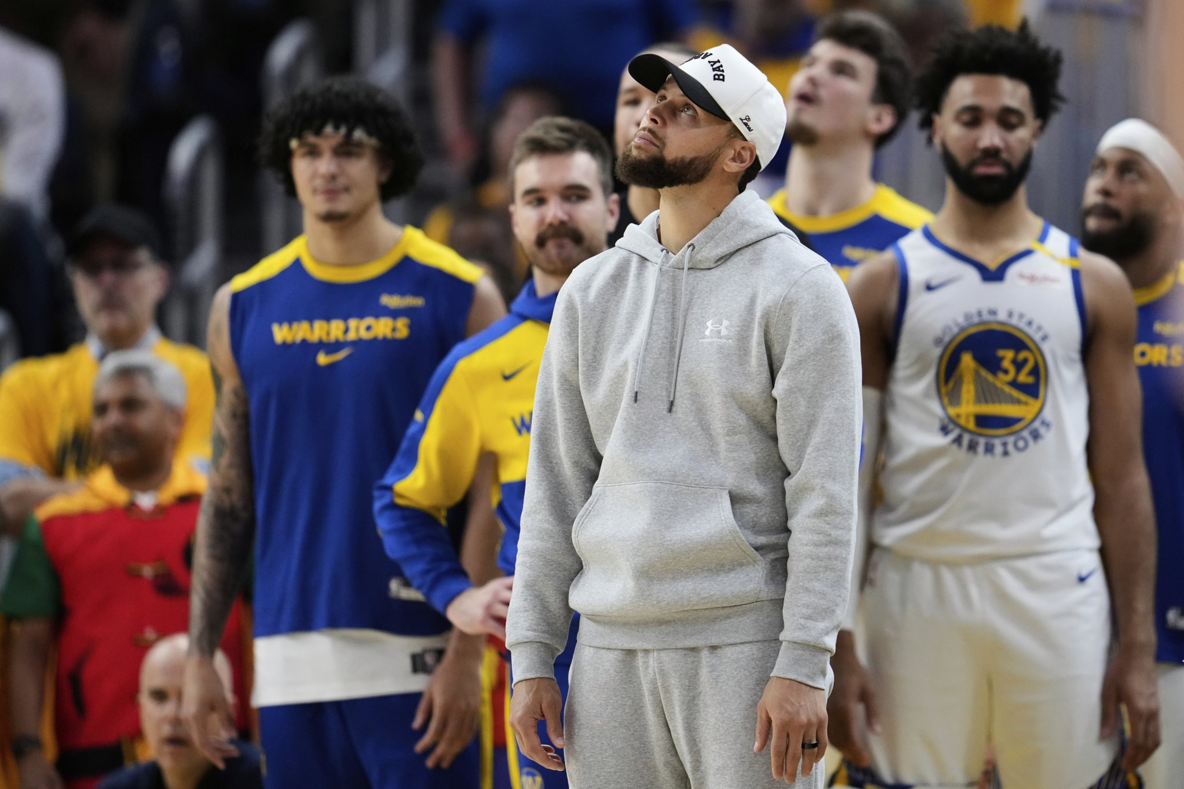 A person in a gray hoodie and white cap stands in front of basketball players wearing Golden State Warriors uniforms during a game.