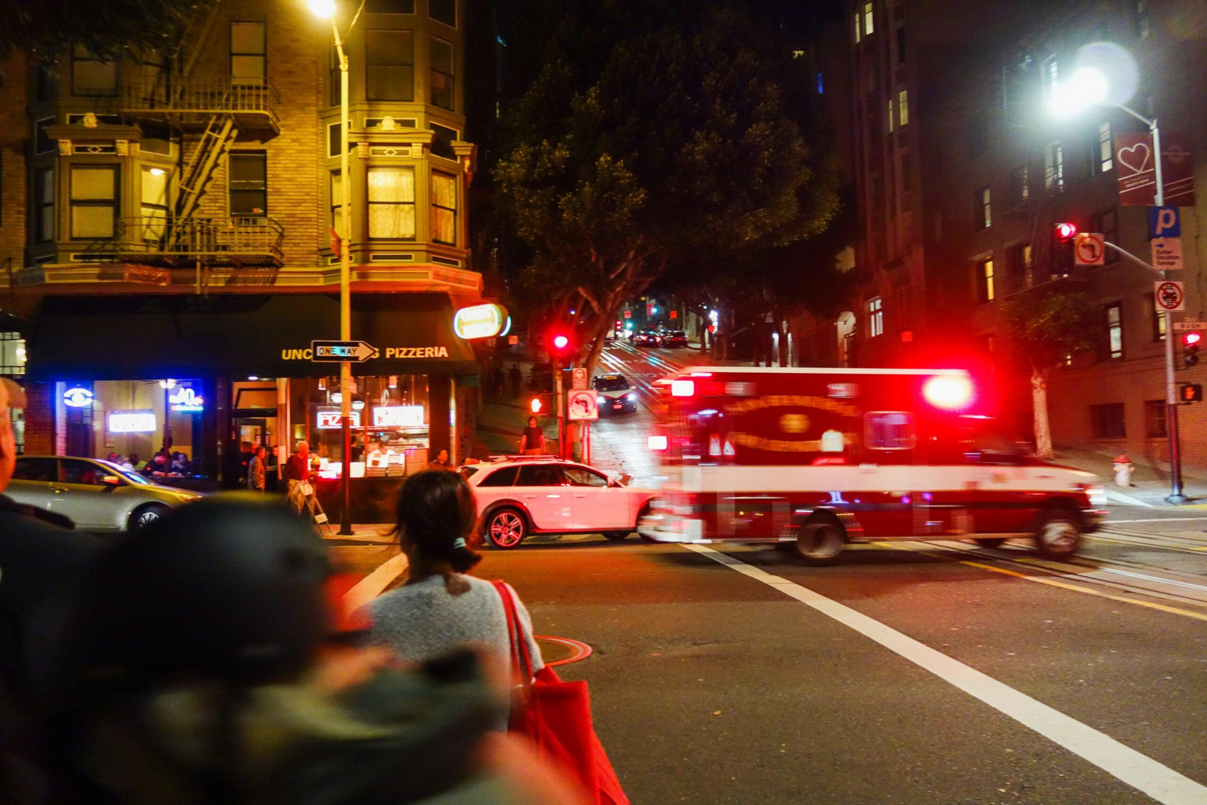 An ambulance with flashing red lights speeds through an intersection at night near a lit pizzeria, while people wait on the sidewalk.