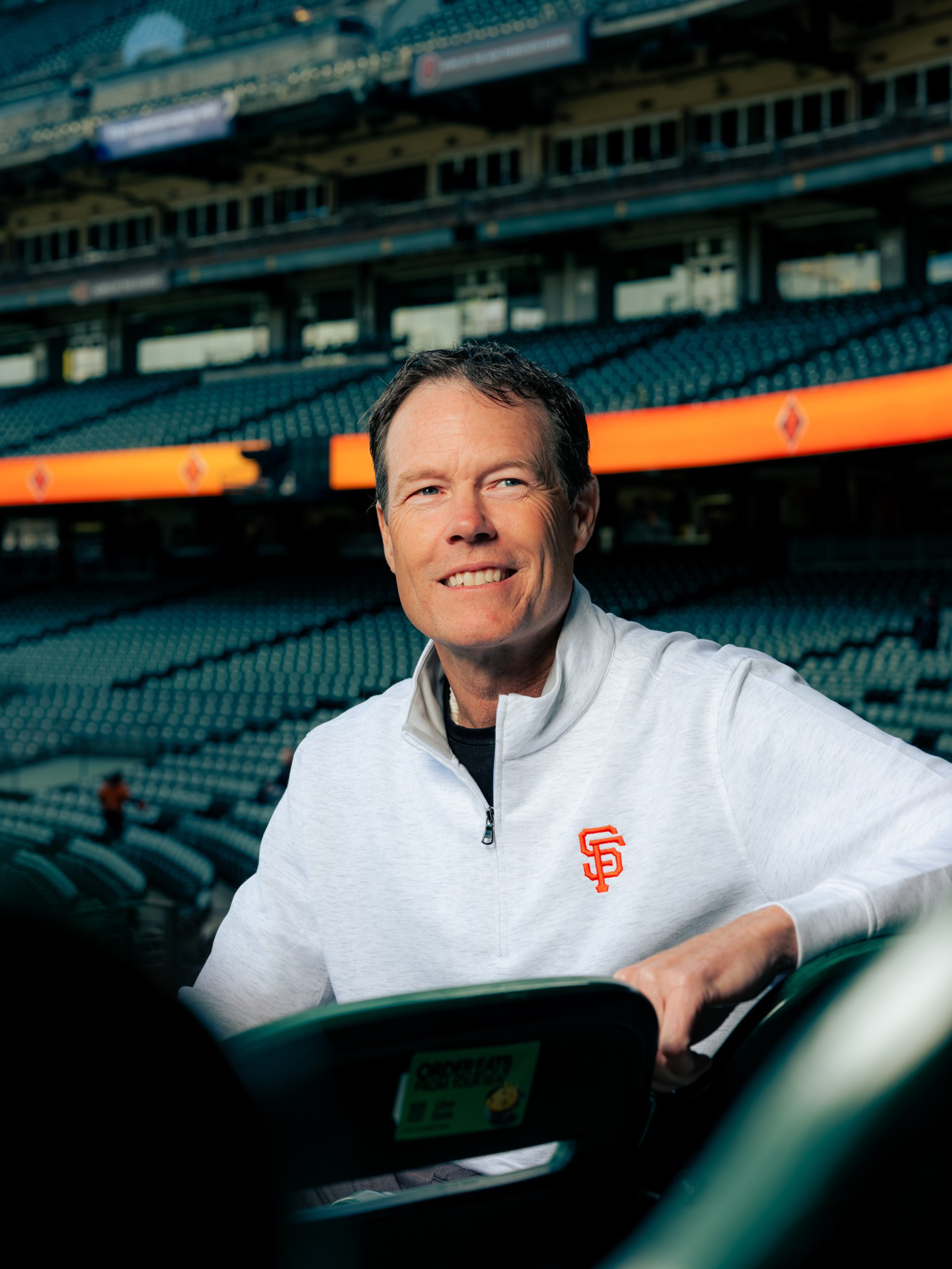 A smiling man in a white jacket with the San Francisco Giants logo sits in empty stadium seats with an orange LED banner in the background.