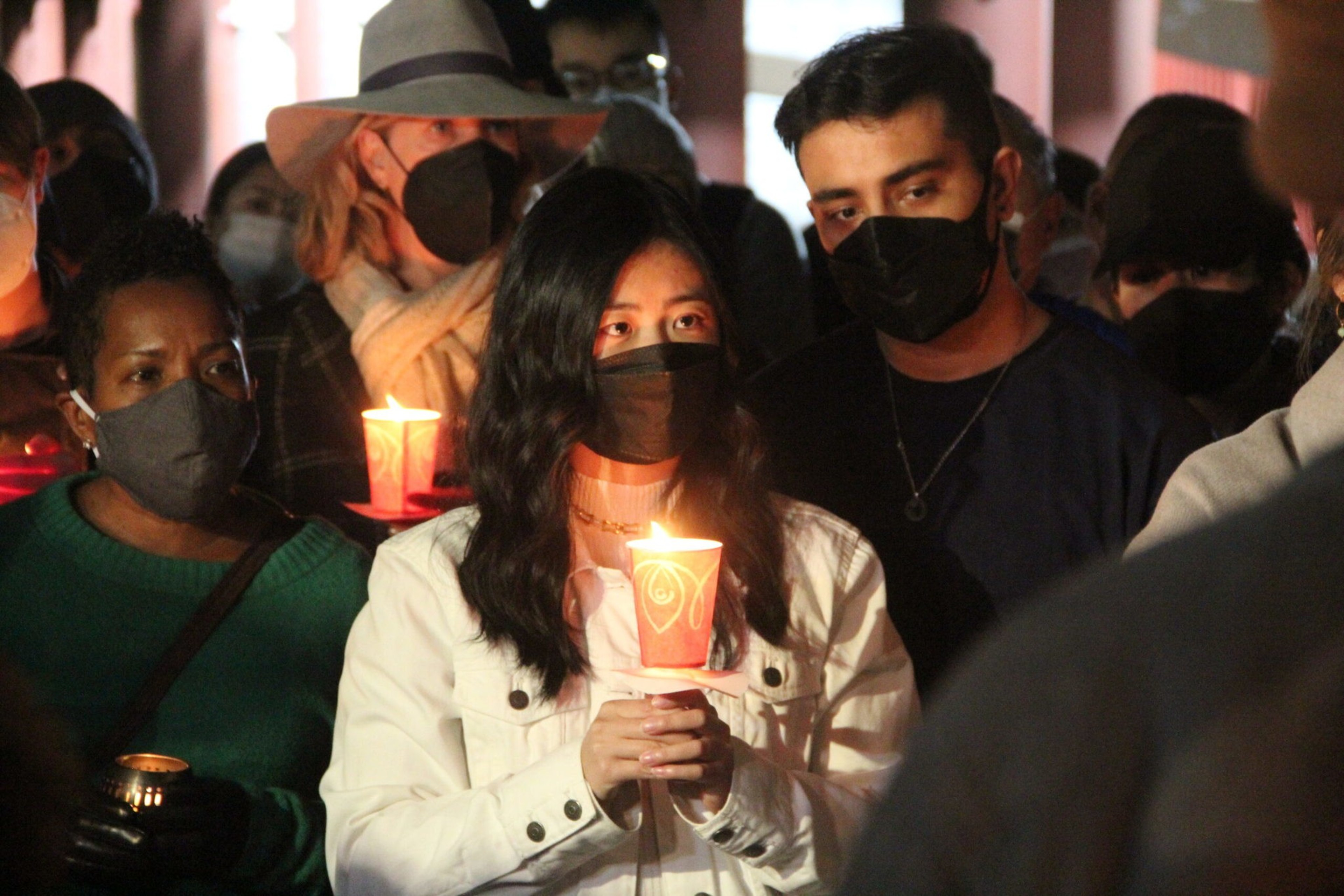 A group of people wearing face masks holds lit candles during a nighttime vigil, their faces illuminated by the candlelight.