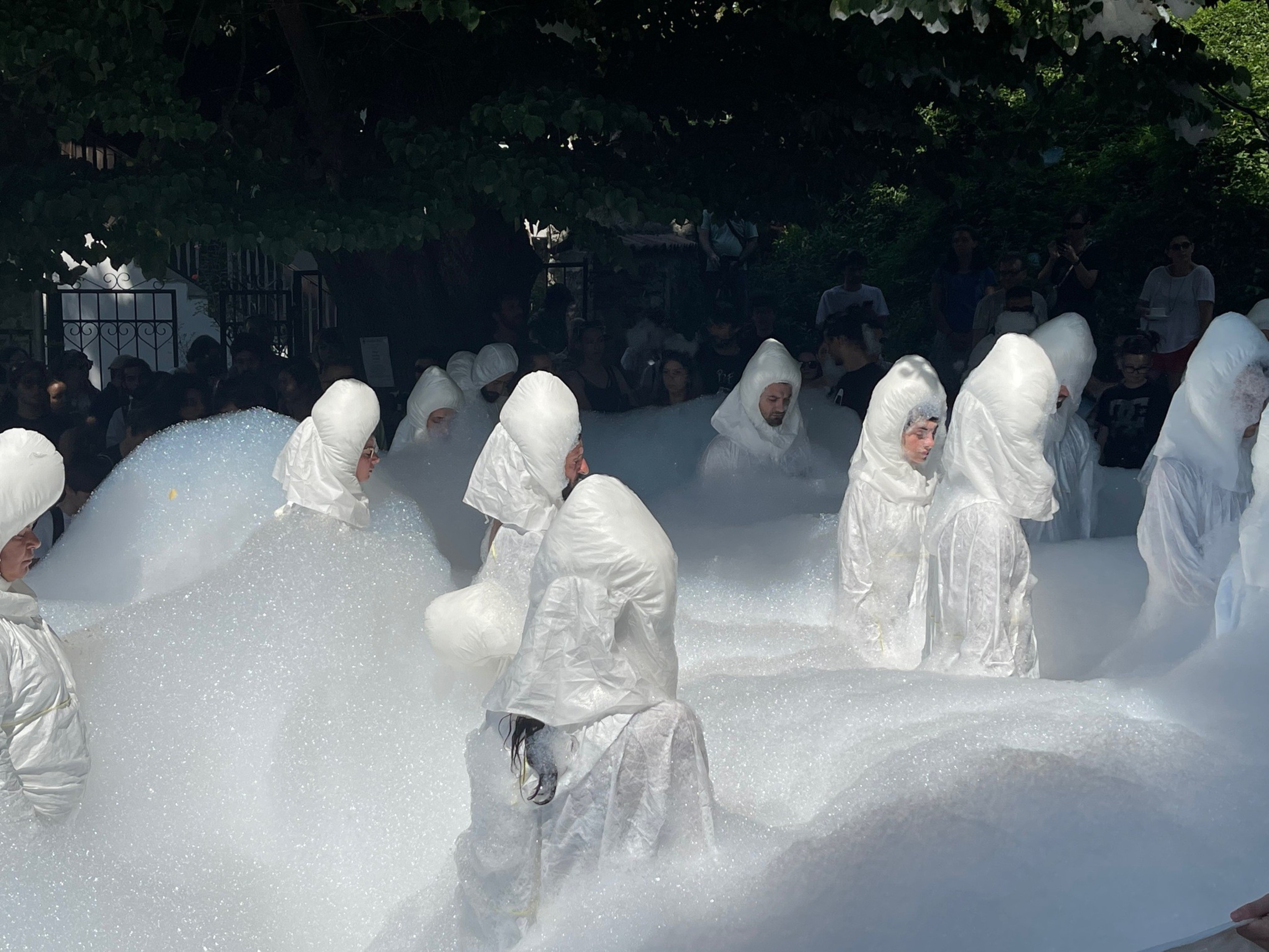 Several people in white hooded outfits stand in thick white foam, surrounded by a crowd under trees, creating a surreal, bubble-filled scene.