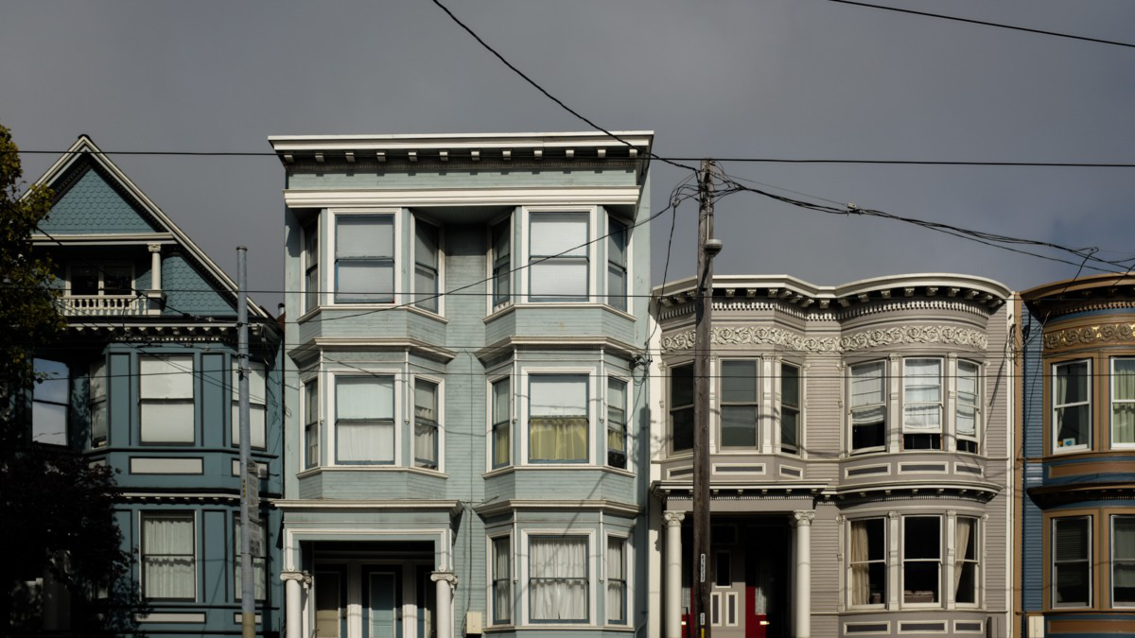 Three Victorian-style houses with bay windows stand side by side under a gray sky, with power lines crossing above.