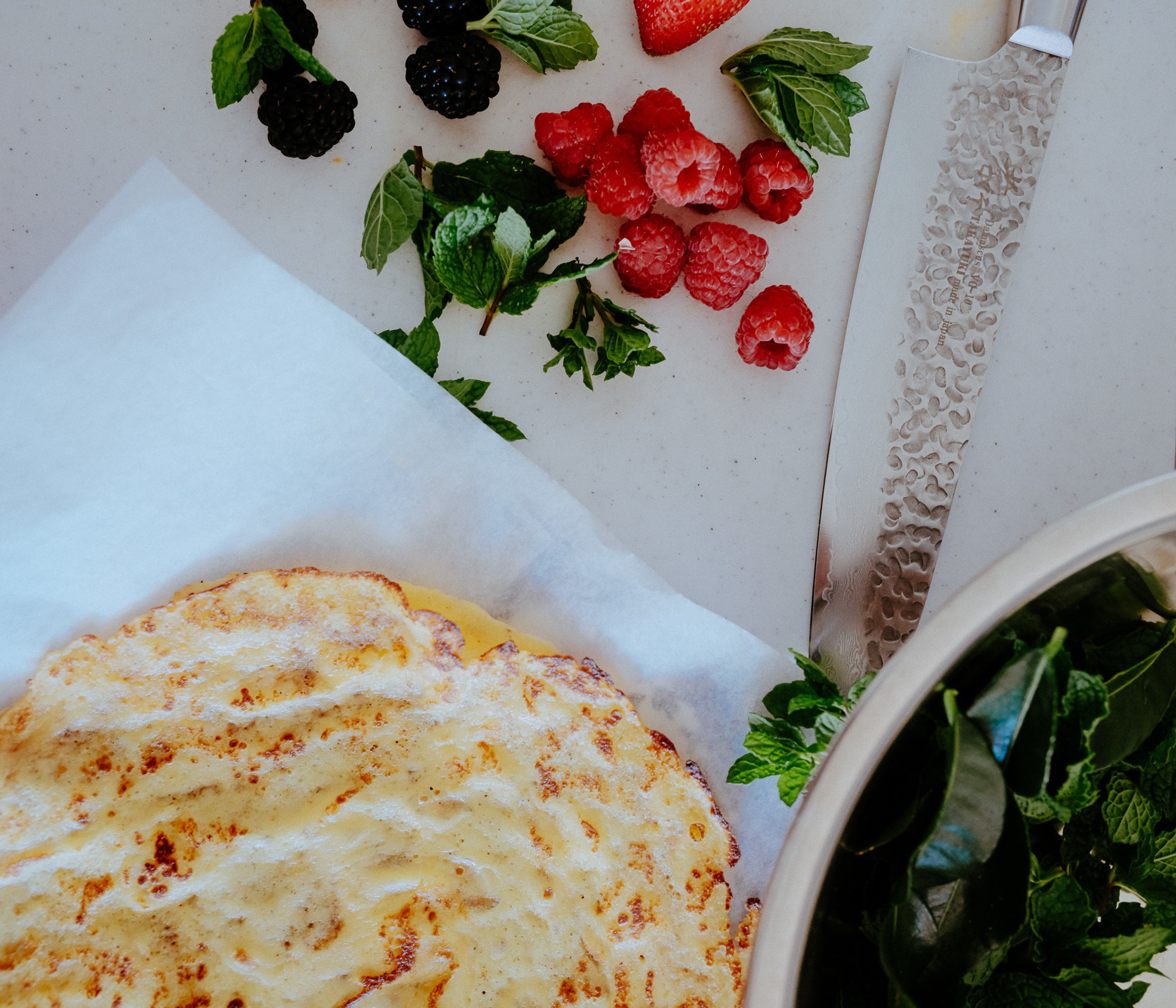 A golden, cooked flatbread rests on white parchment paper, surrounded by fresh raspberries, blackberries, mint leaves, a large textured knife, and a bowl of greens.