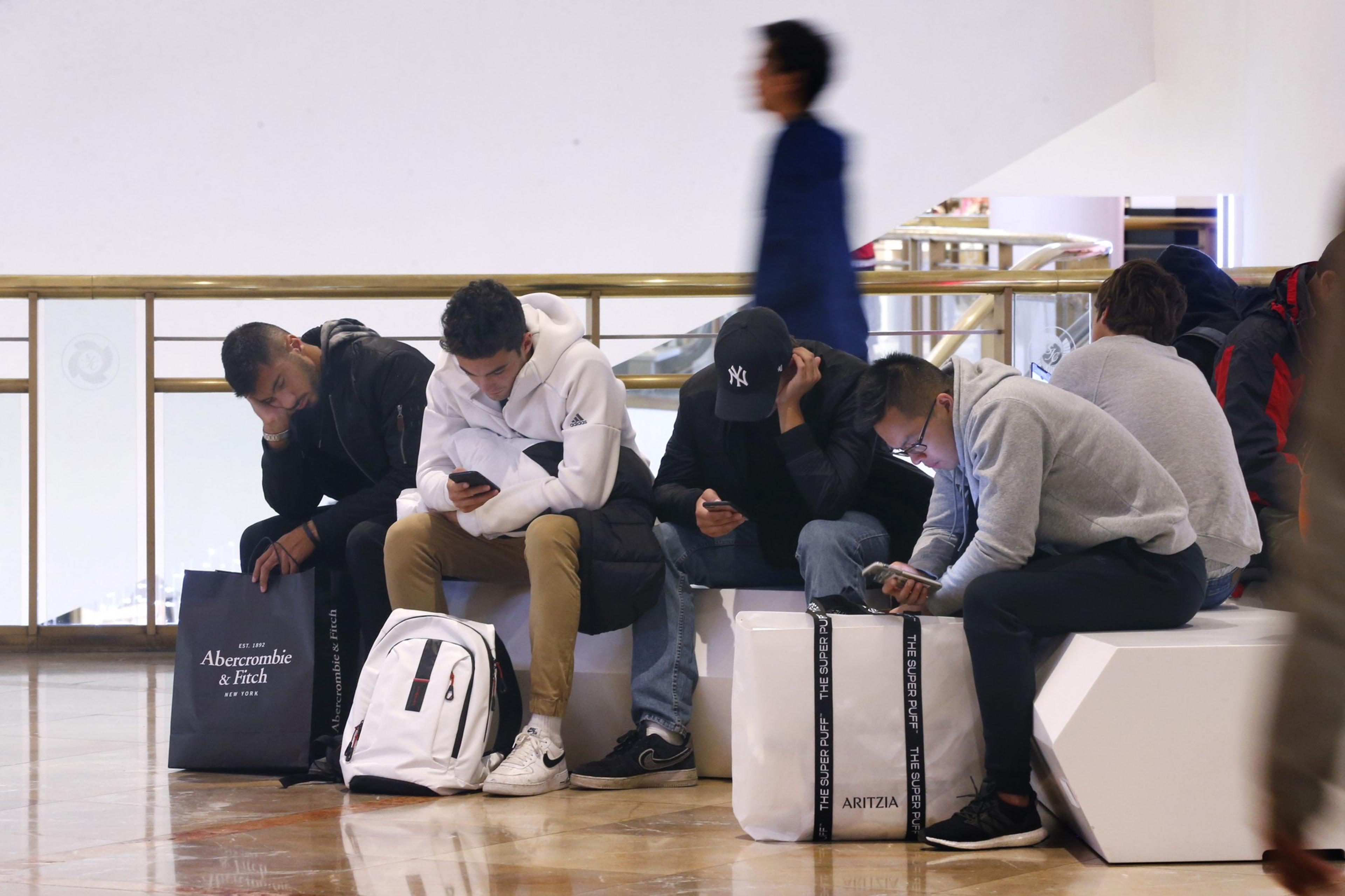 People sit on a bench engrossed in their phones, a blurred figure walks by, shopping bags at their feet.