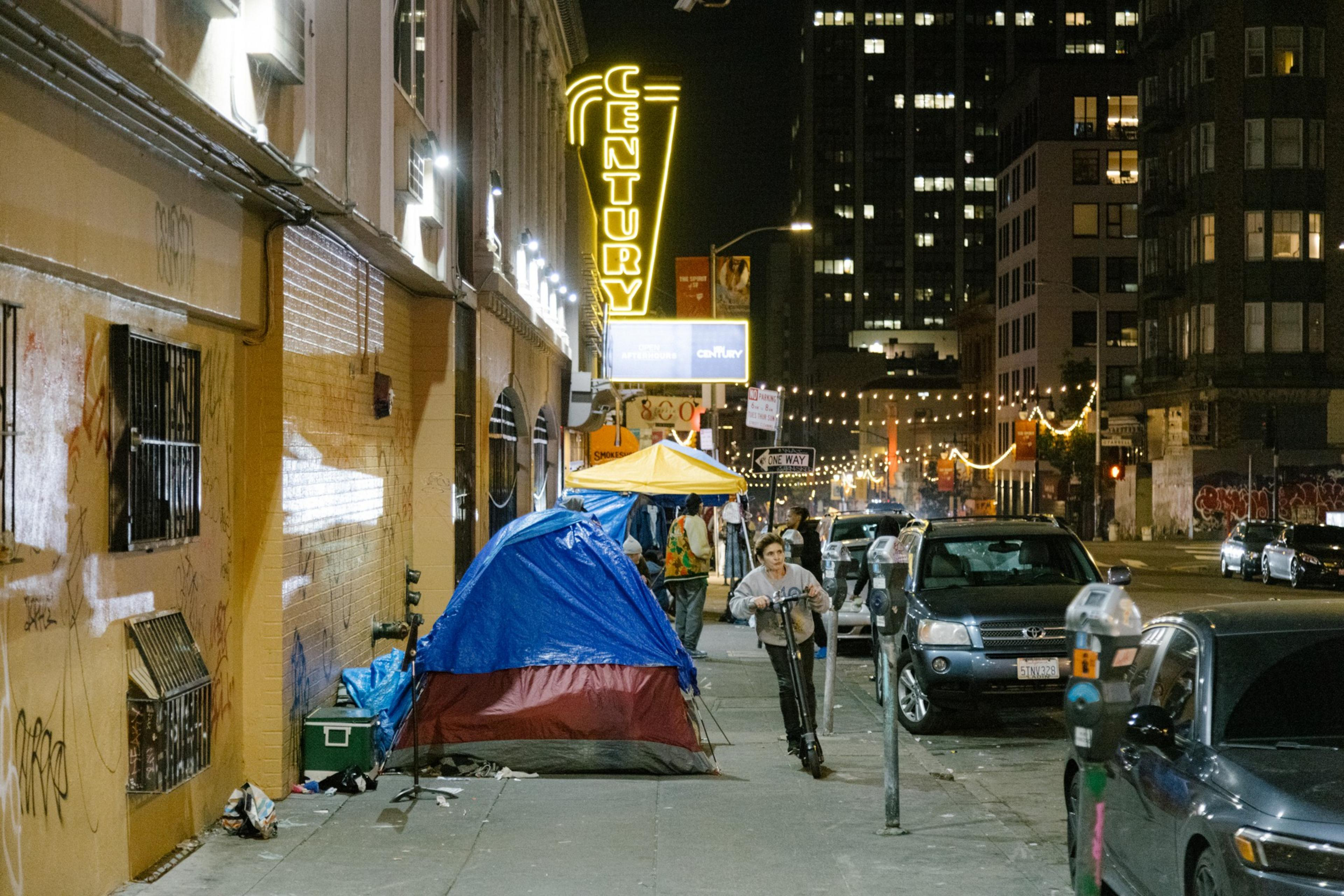 An urban street at night with tents, neon signs, cars, and a person on a scooter.