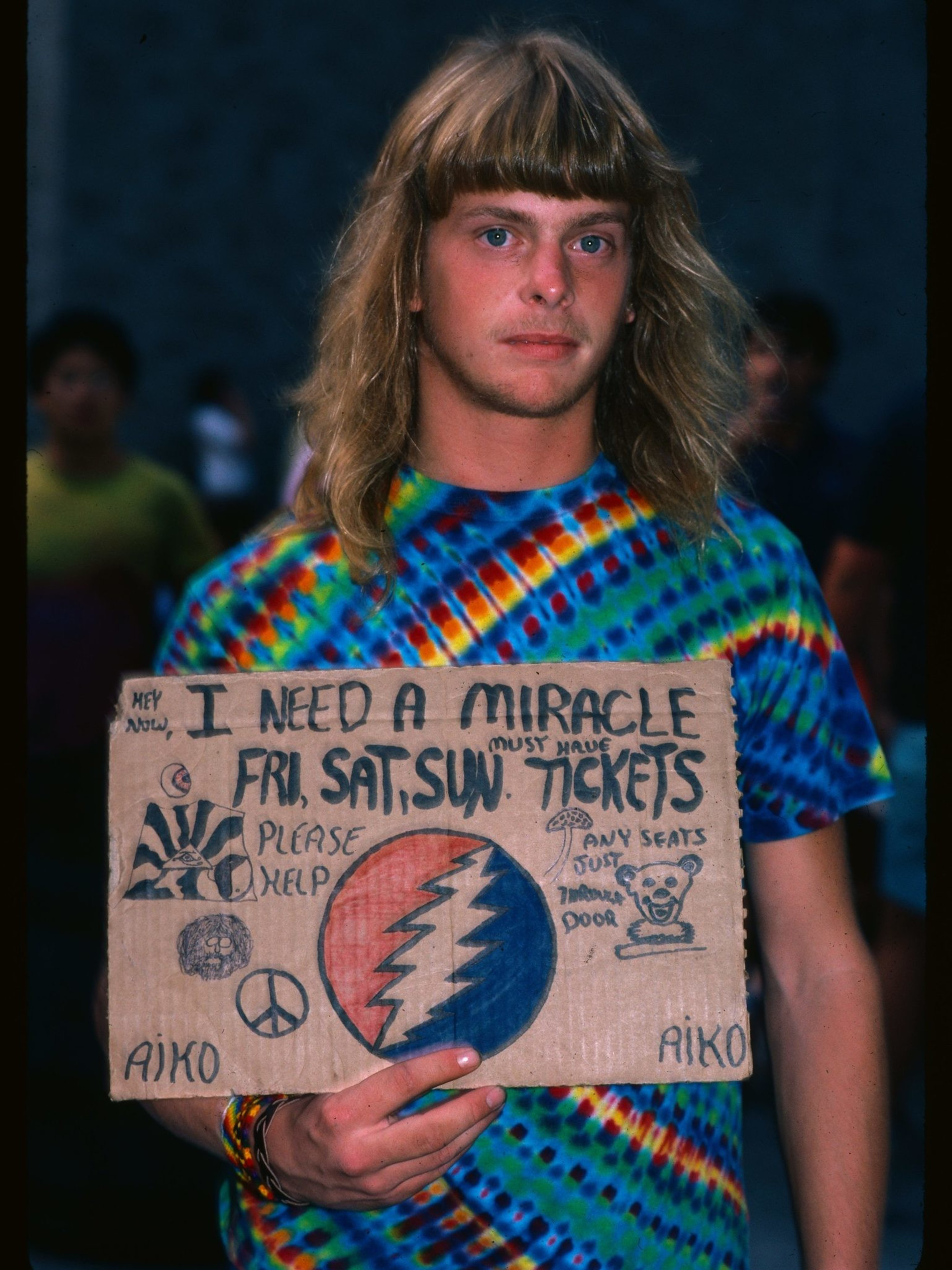 The image shows a person with long blond hair wearing a tie-dye shirt, holding a cardboard sign asking for tickets. The sign includes doodles and peace symbols.