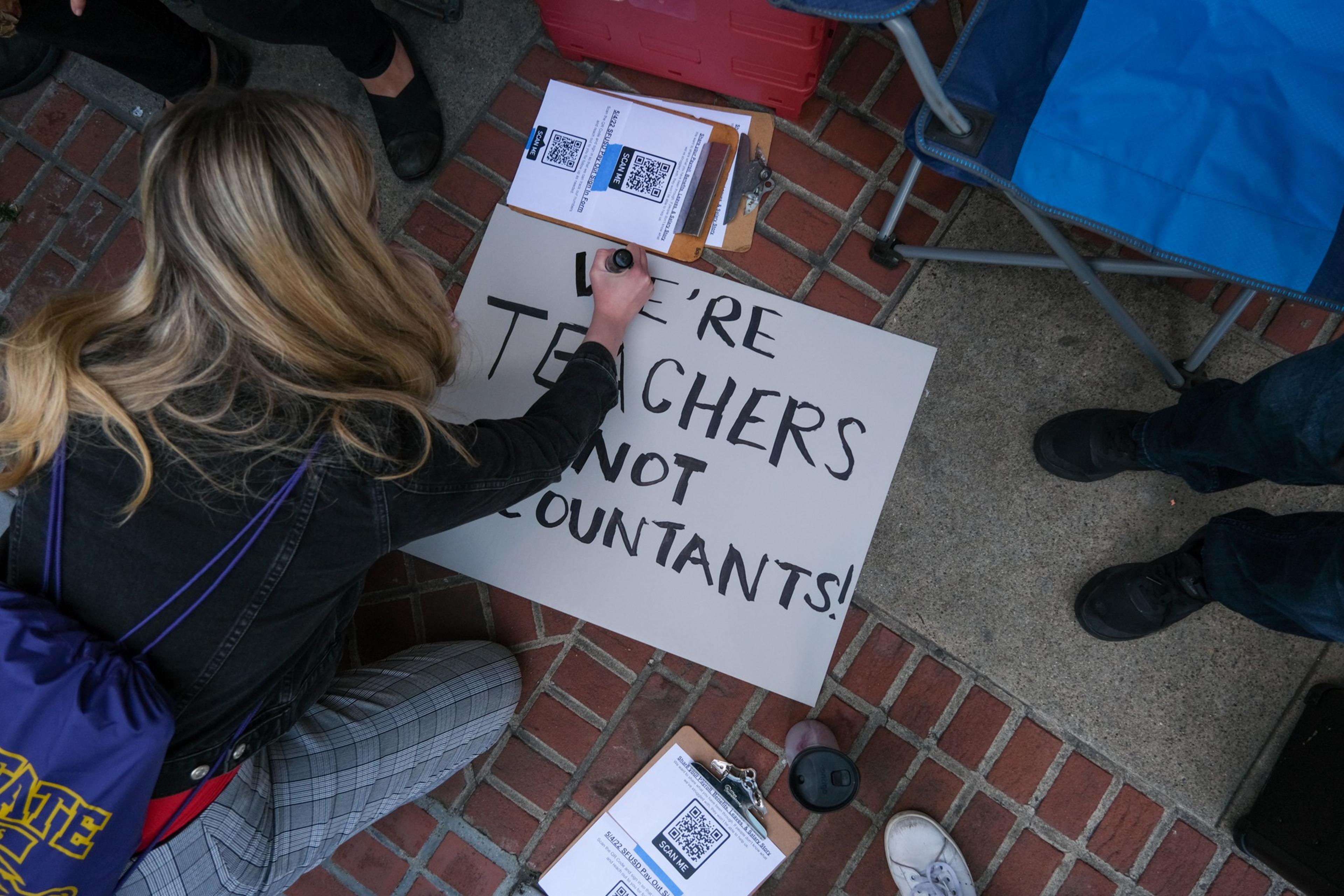 A person with blonde hair writes on a sign that says "WE'RE TEACHERS NOT ACCOUNTANTS!" while sitting on a brick floor near clipboards and people.