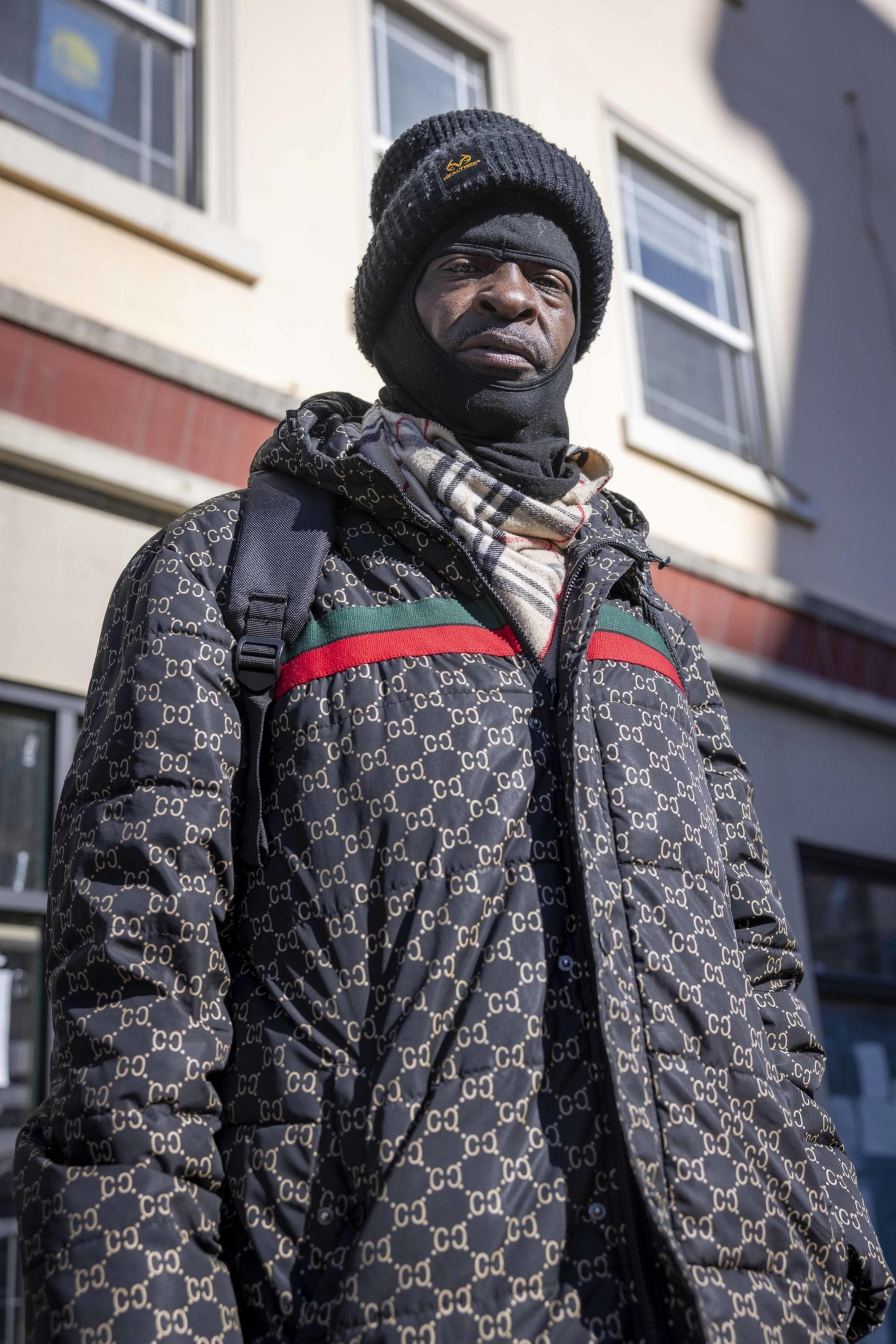 A person stands outside a building wearing a patterned winter coat, a black beanie, a balaclava, and a plaid scarf. Sunlight casts shadows on the wall.