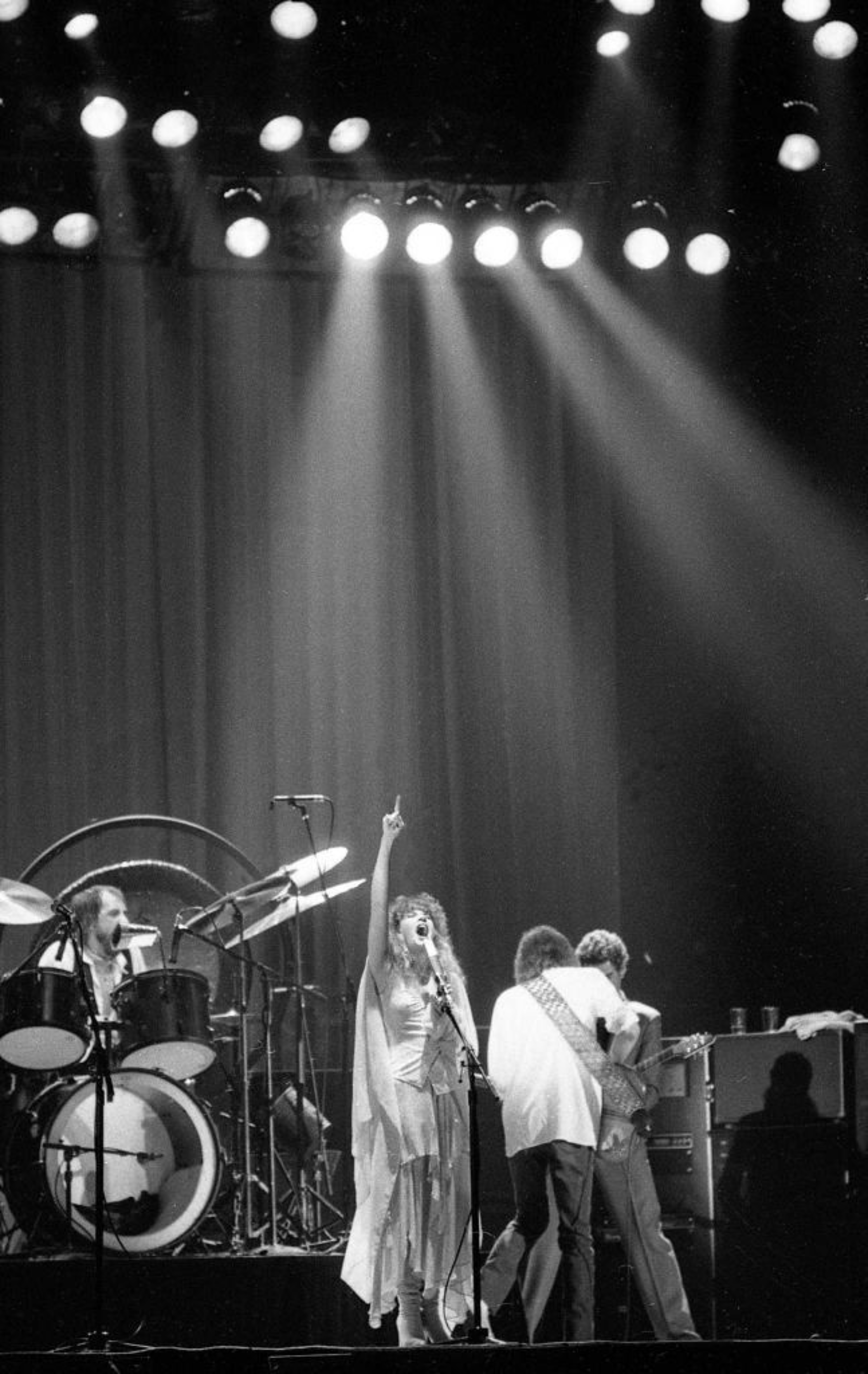 A black-and-white photo of a band on stage. A singer gestures upward, with musicians and stage lights around her.
