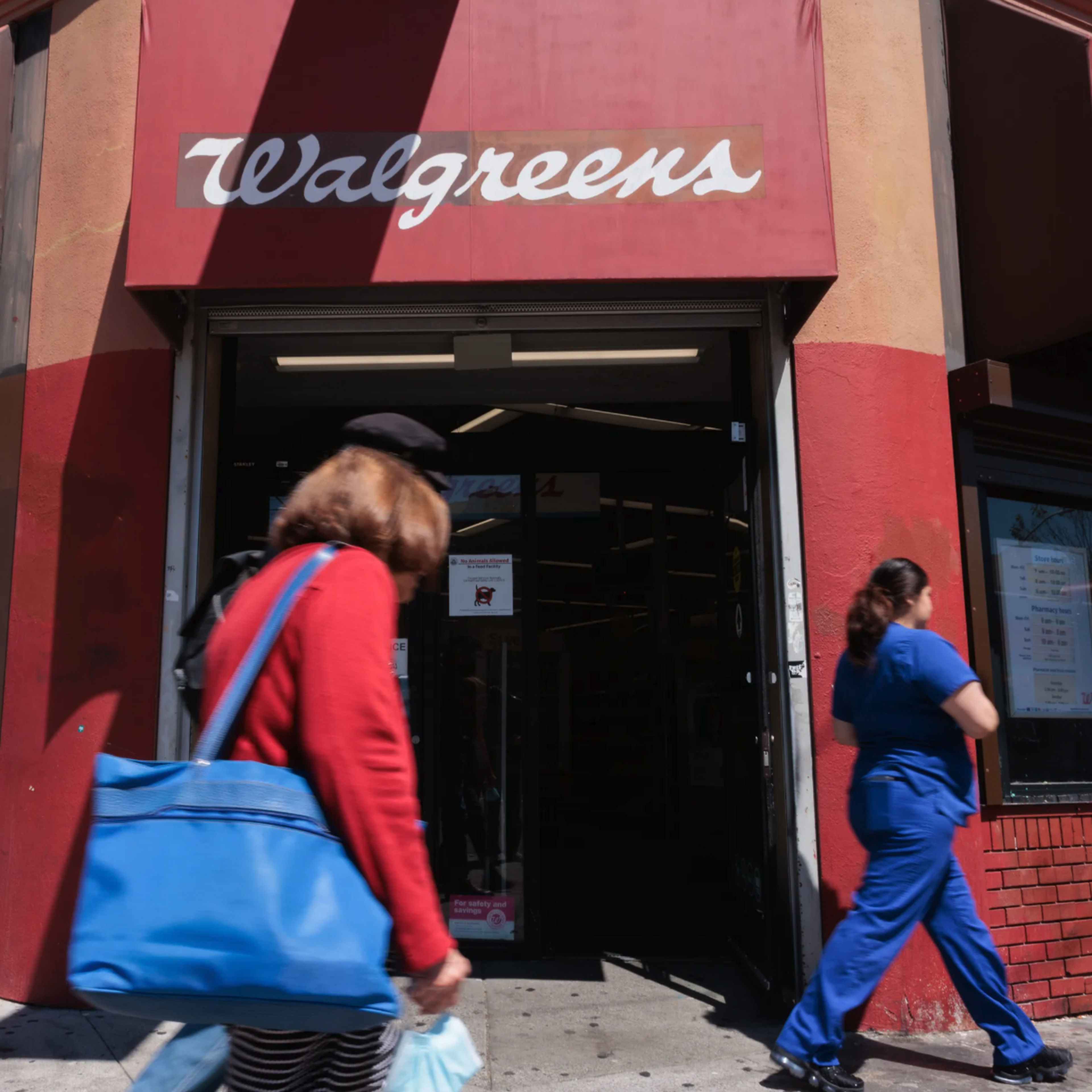 A storefront with a Walgreens sign, featuring a brick facade. Two people are walking by, one in a red coat holding a blue bag, and the other in blue scrubs.