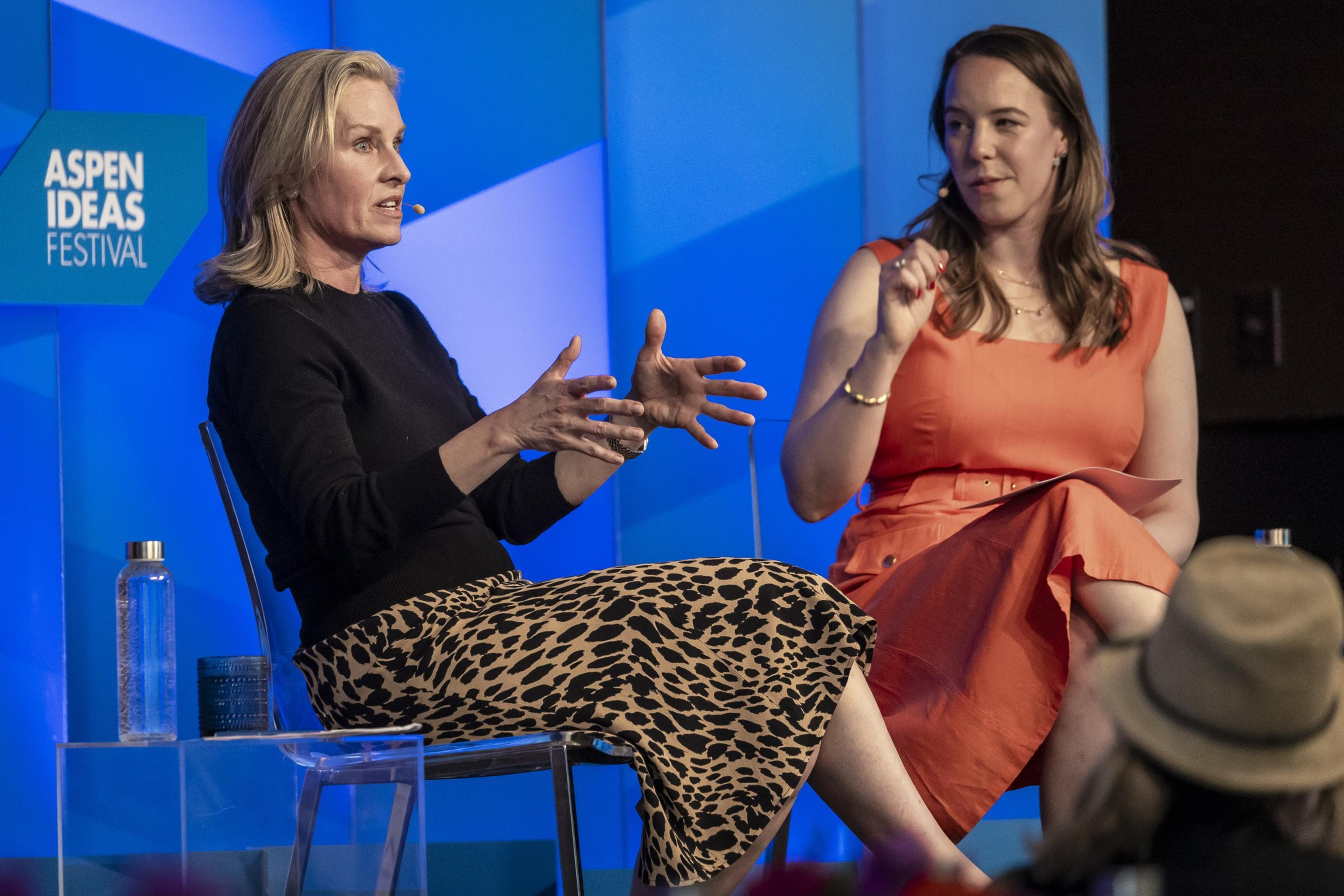 Two women are seated and discussing on stage at the Aspen Ideas Festival; one gestures with hands while the other listens attentively in an orange dress.