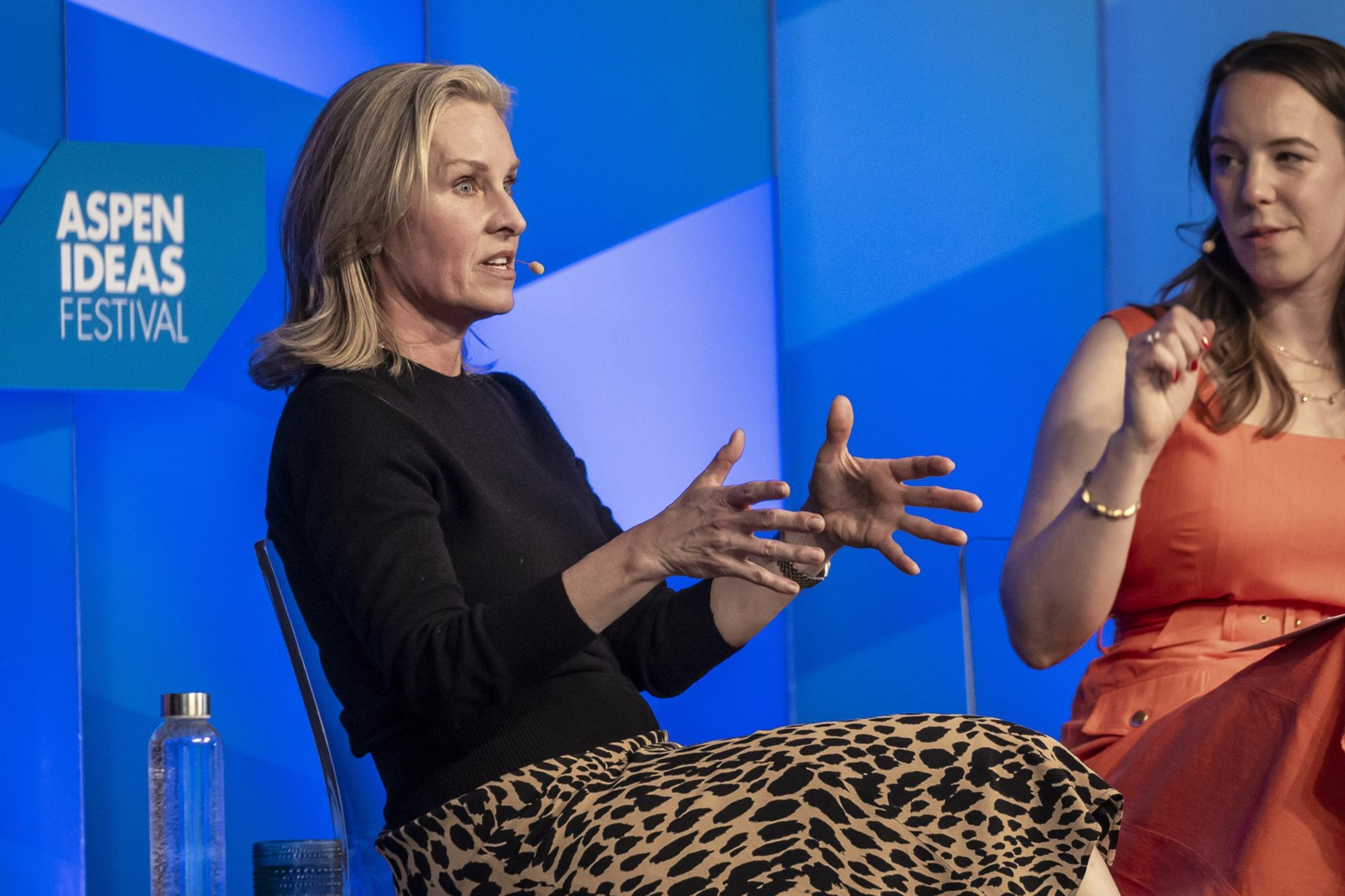 Two women are seated and discussing on stage at the Aspen Ideas Festival; one gestures with hands while the other listens attentively in an orange dress.