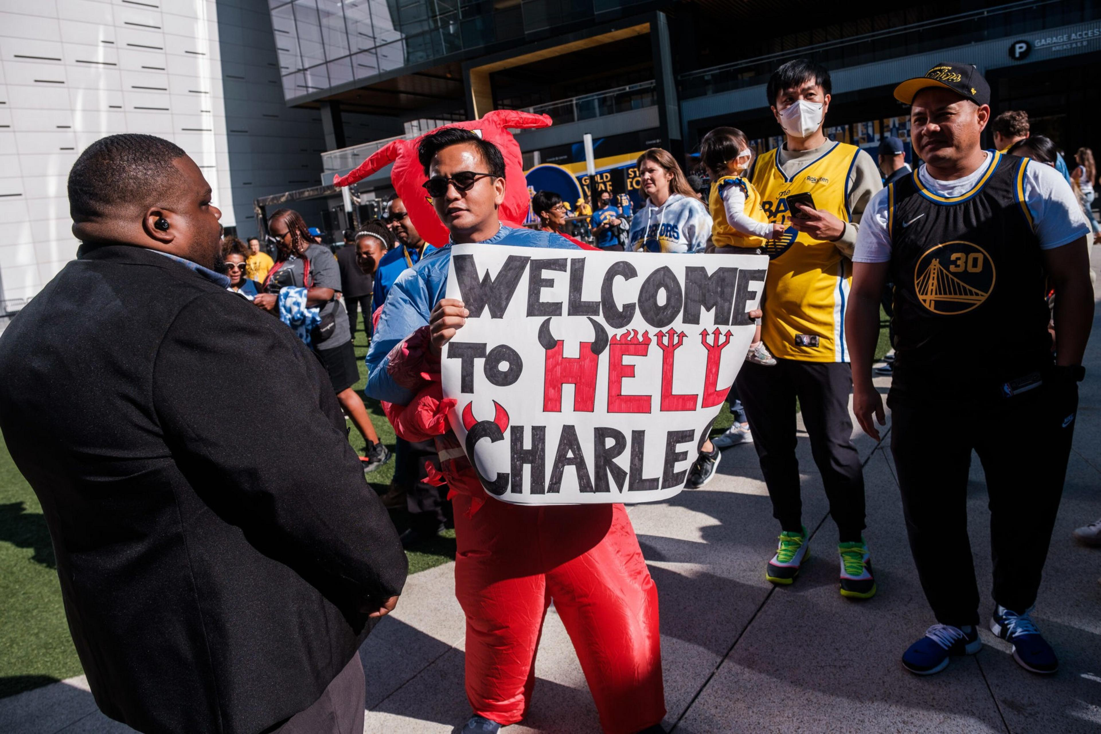 A man in red stands holding a sign saying &quot;Welcome to Hell, Charles.&quot; He's surrounded by people, including some in sports jerseys, outside a modern building.