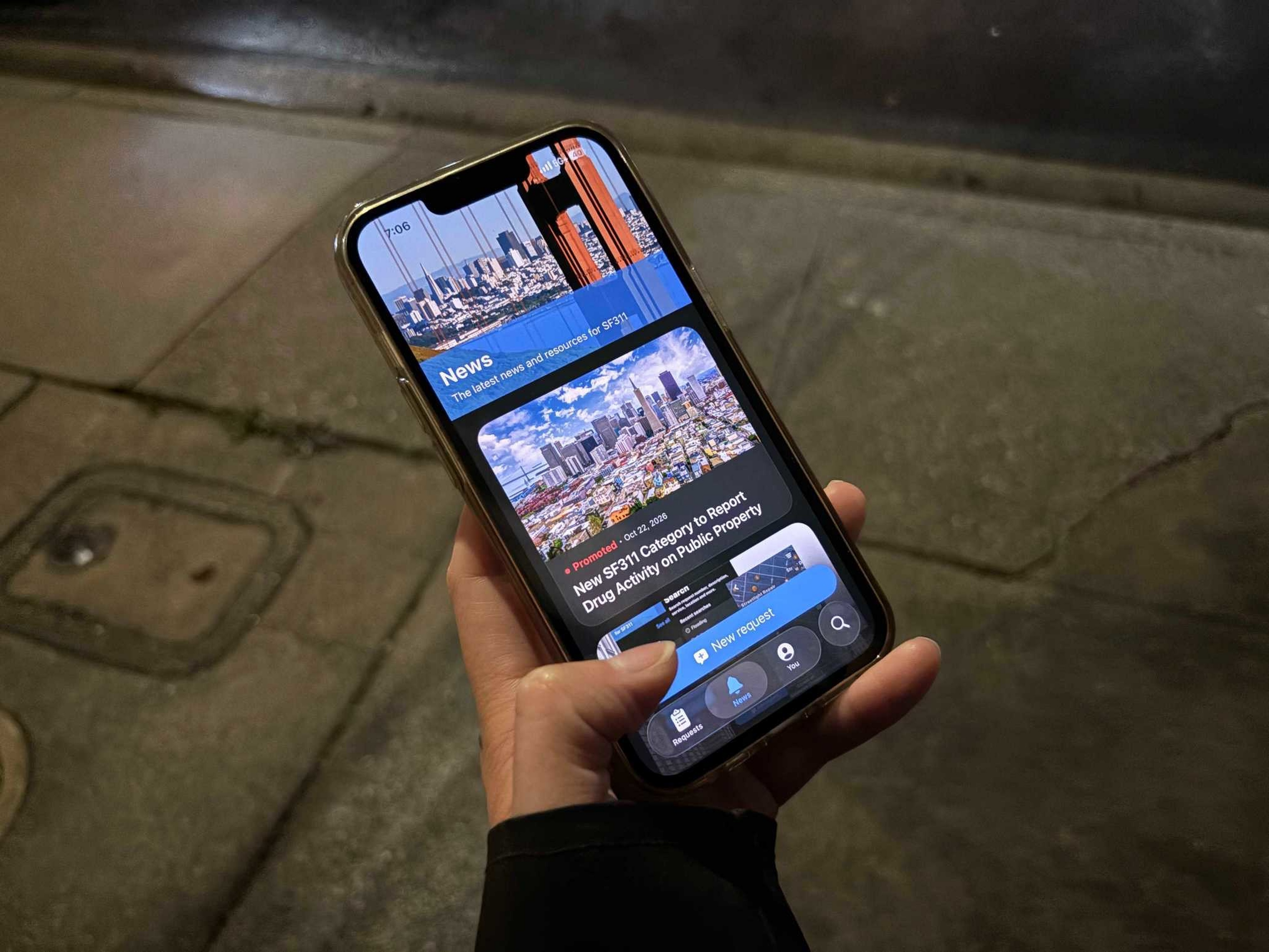 A hand holds a smartphone displaying a news app with articles and images about San Francisco, including a report on drug activity in public areas.