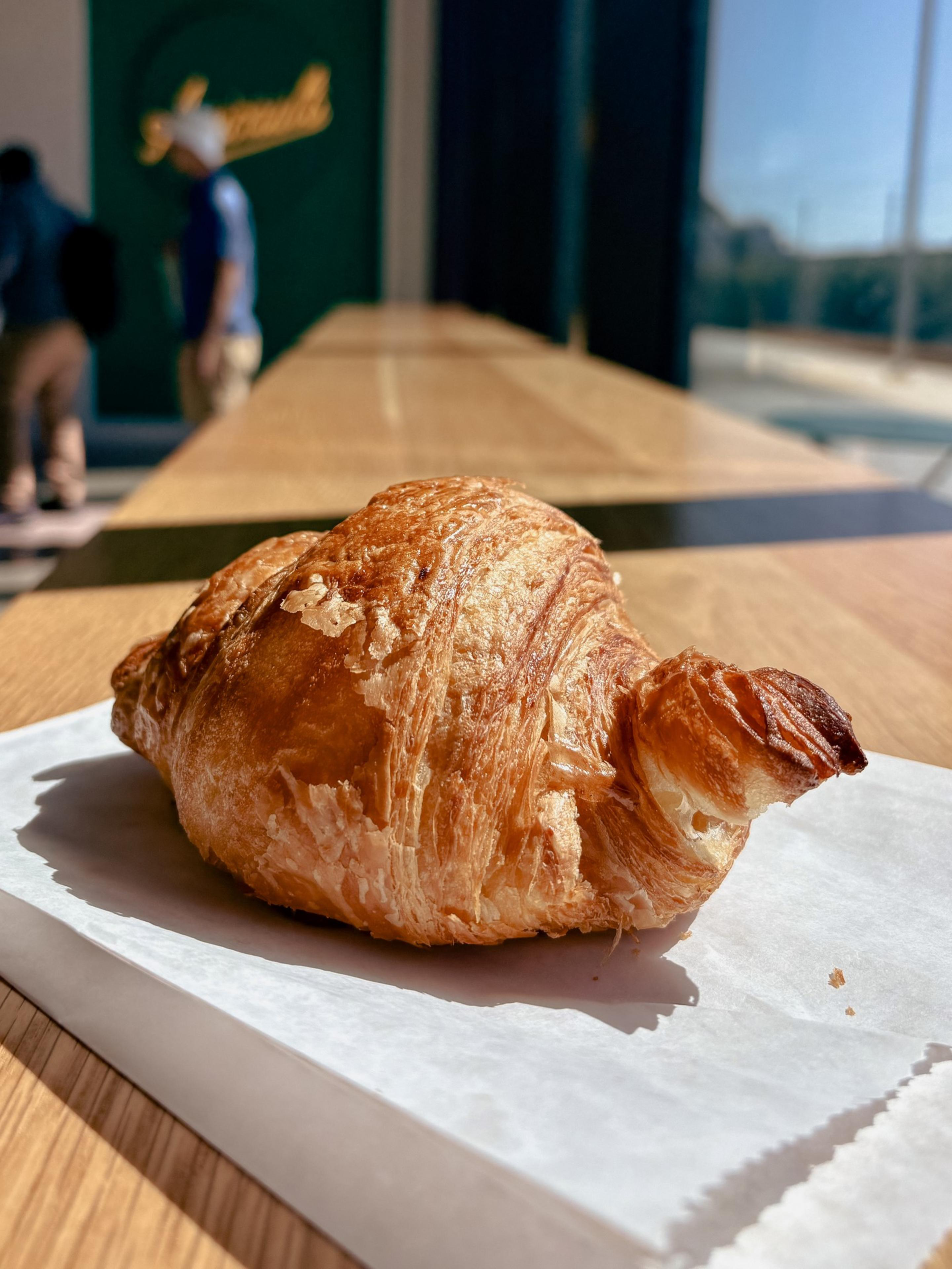 A golden-brown, flaky croissant rests on white parchment paper atop a wooden table, with blurred figures and sunlight in the background.