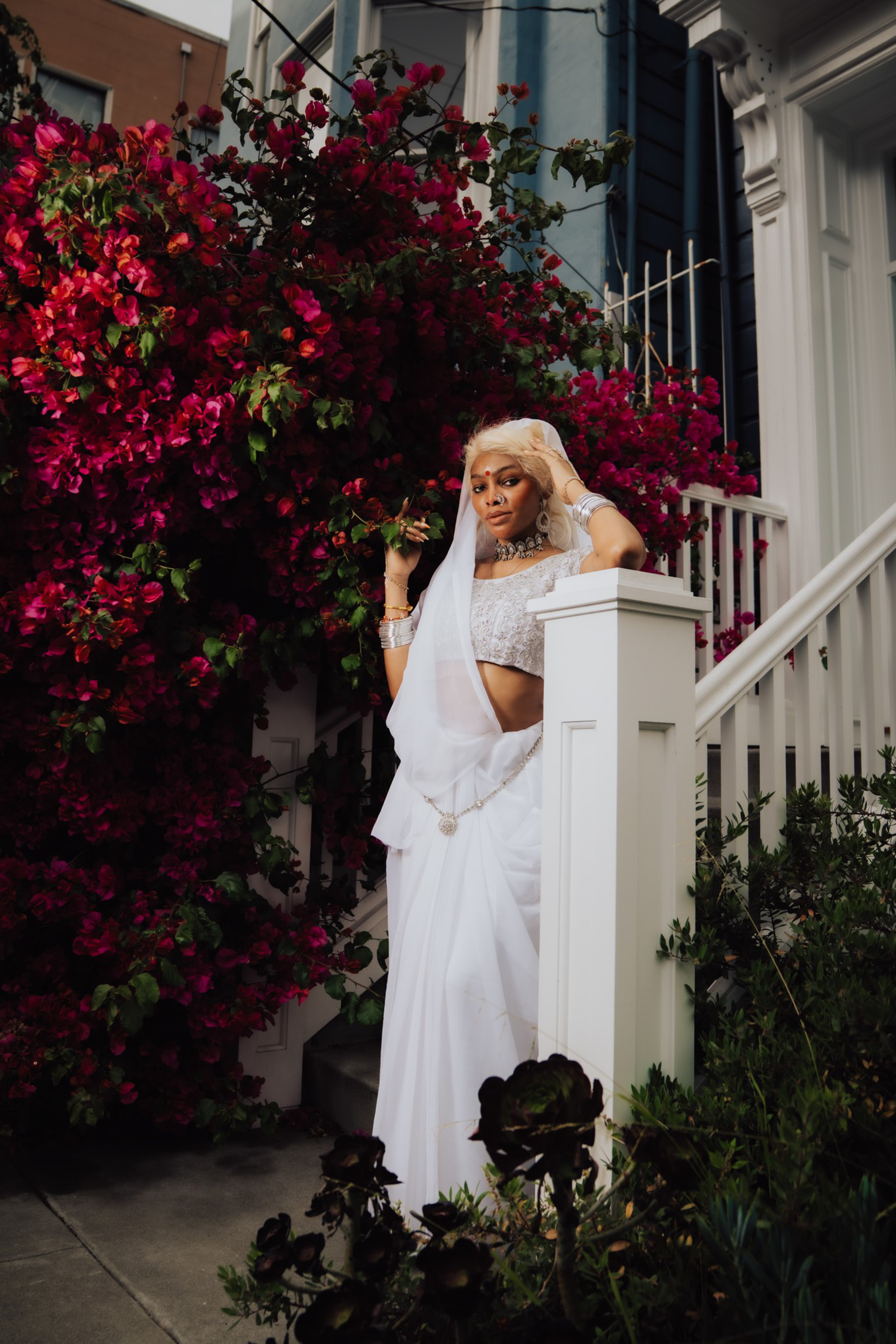 A woman in a white sari and silver jewelry stands by a white railing, surrounded by vibrant red flowers and greenery.