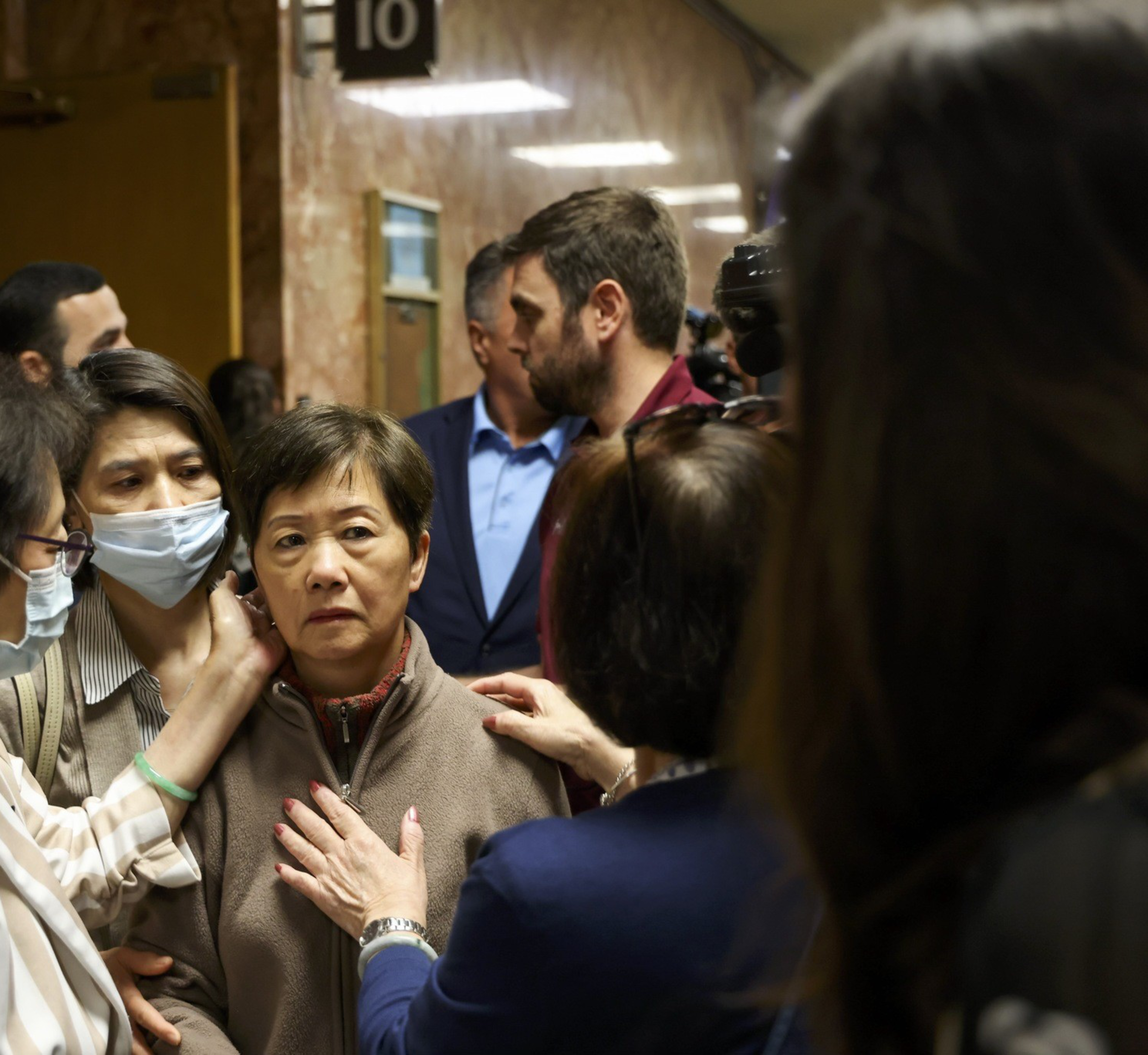A woman wearing a mask that covers her mouth and nose caresses the face of a woman in a crowded hallway outside of a courtroom.