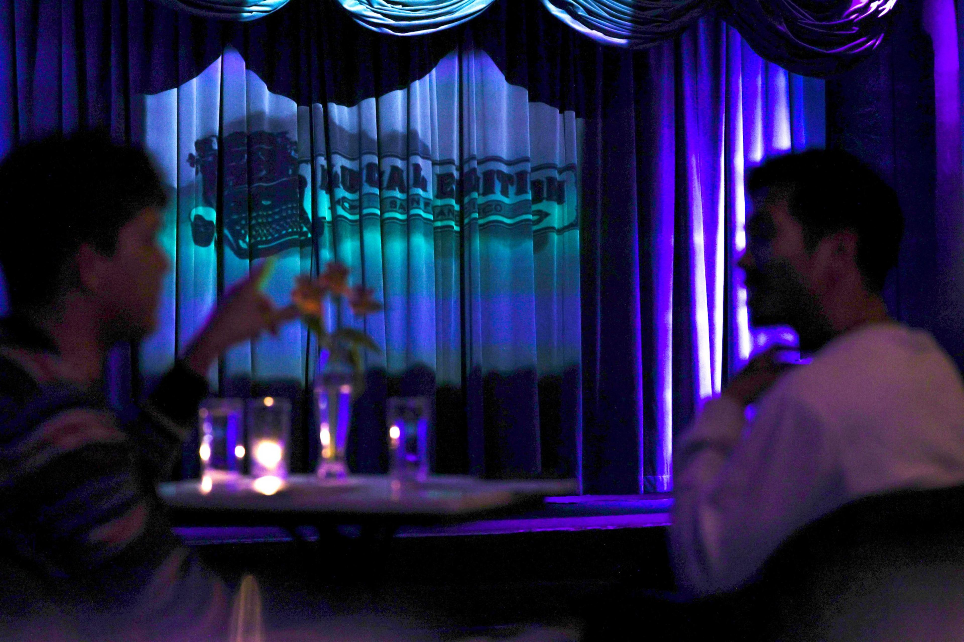 Two people sit in a dimly lit room at a table with candles and flowers. The background features a stage with closed curtains and a typewriter silhouette.