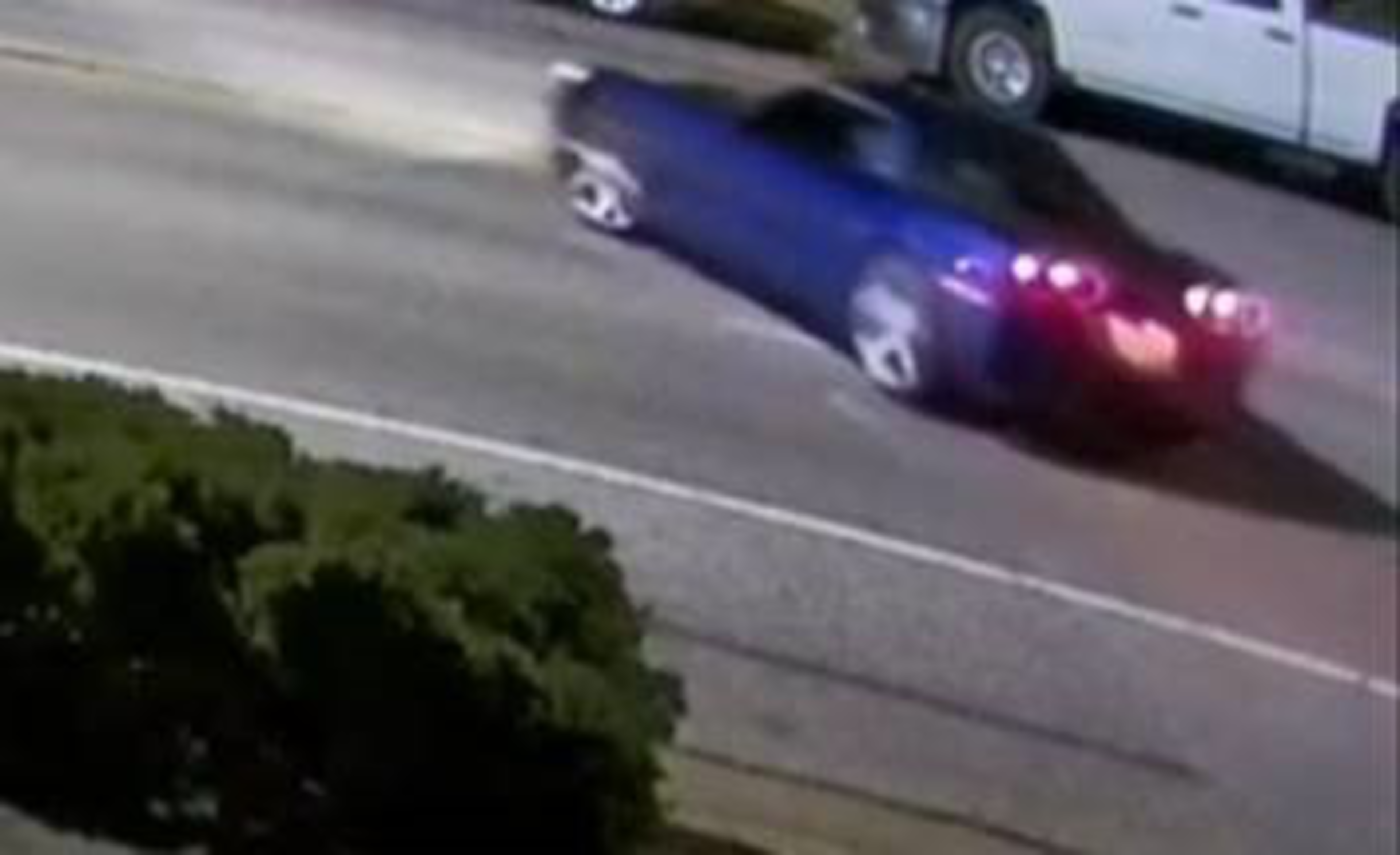 A blue car with its taillights on is driving on a street near a sidewalk with bushes on one side and a white pickup truck parked on the other side.
