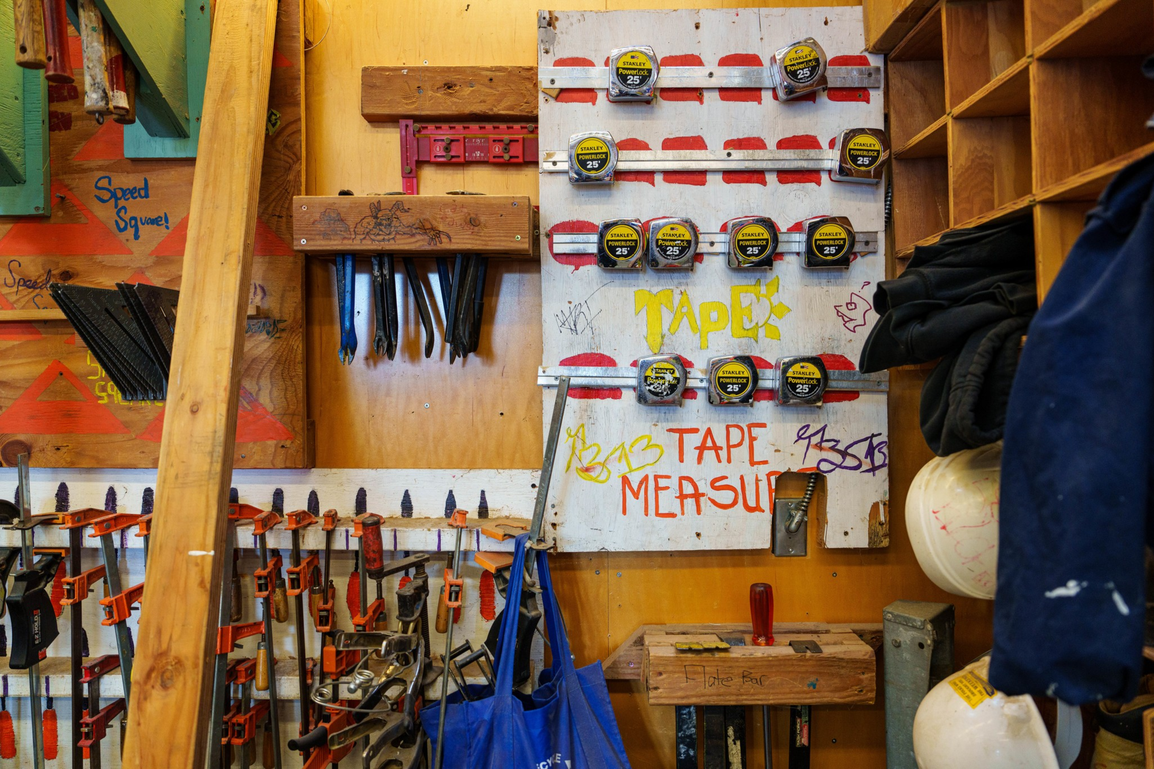 Multiple tape measures are neatly arranged on a white board labeled &ldquo;Tape Measure&rdquo; with various clamps and tools hanging on nearby wooden walls.